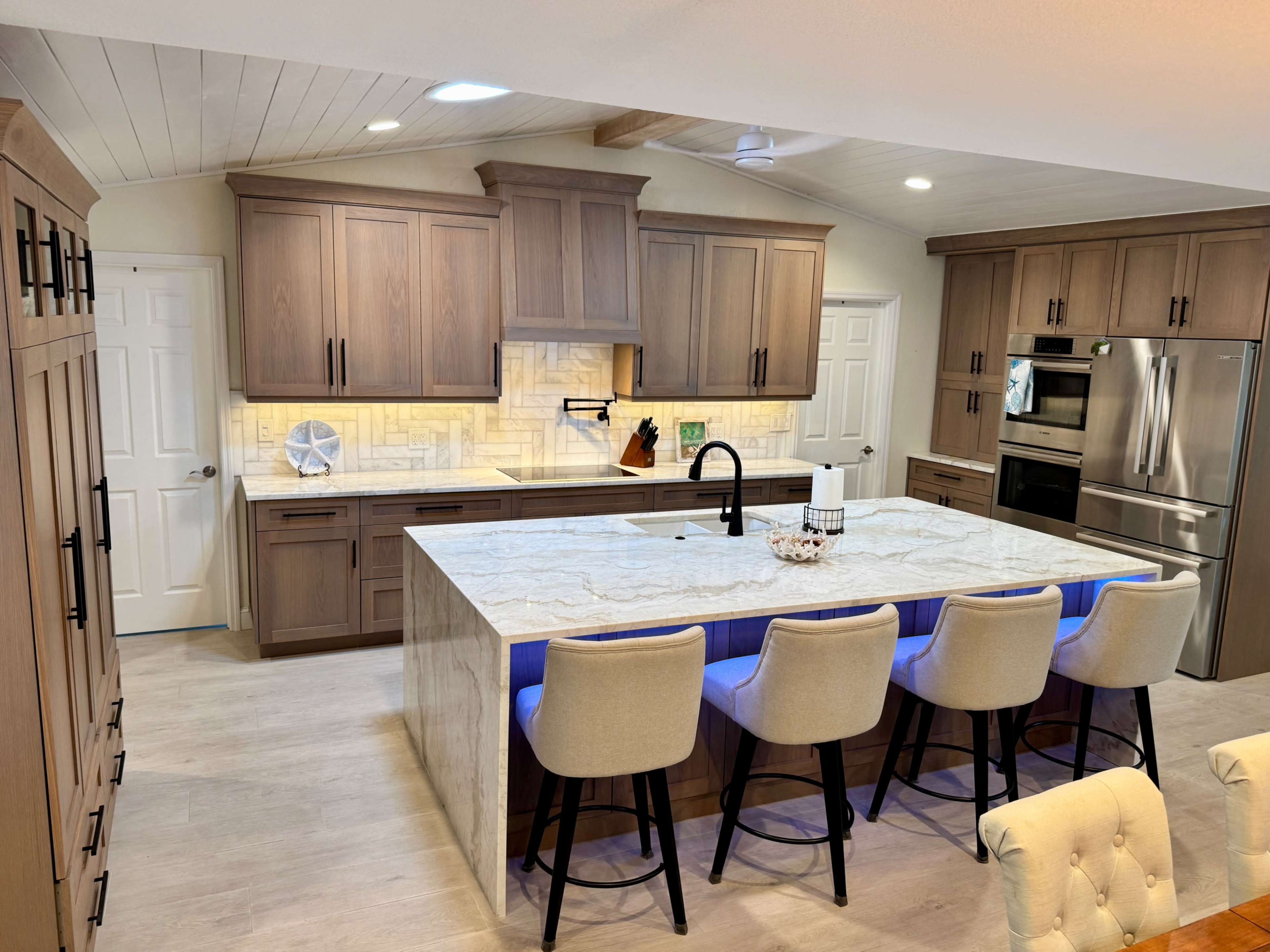 The image shows a modern kitchen featuring wooden cabinetry, a large island with a marble countertop and bar seating, and stainless steel appliances.