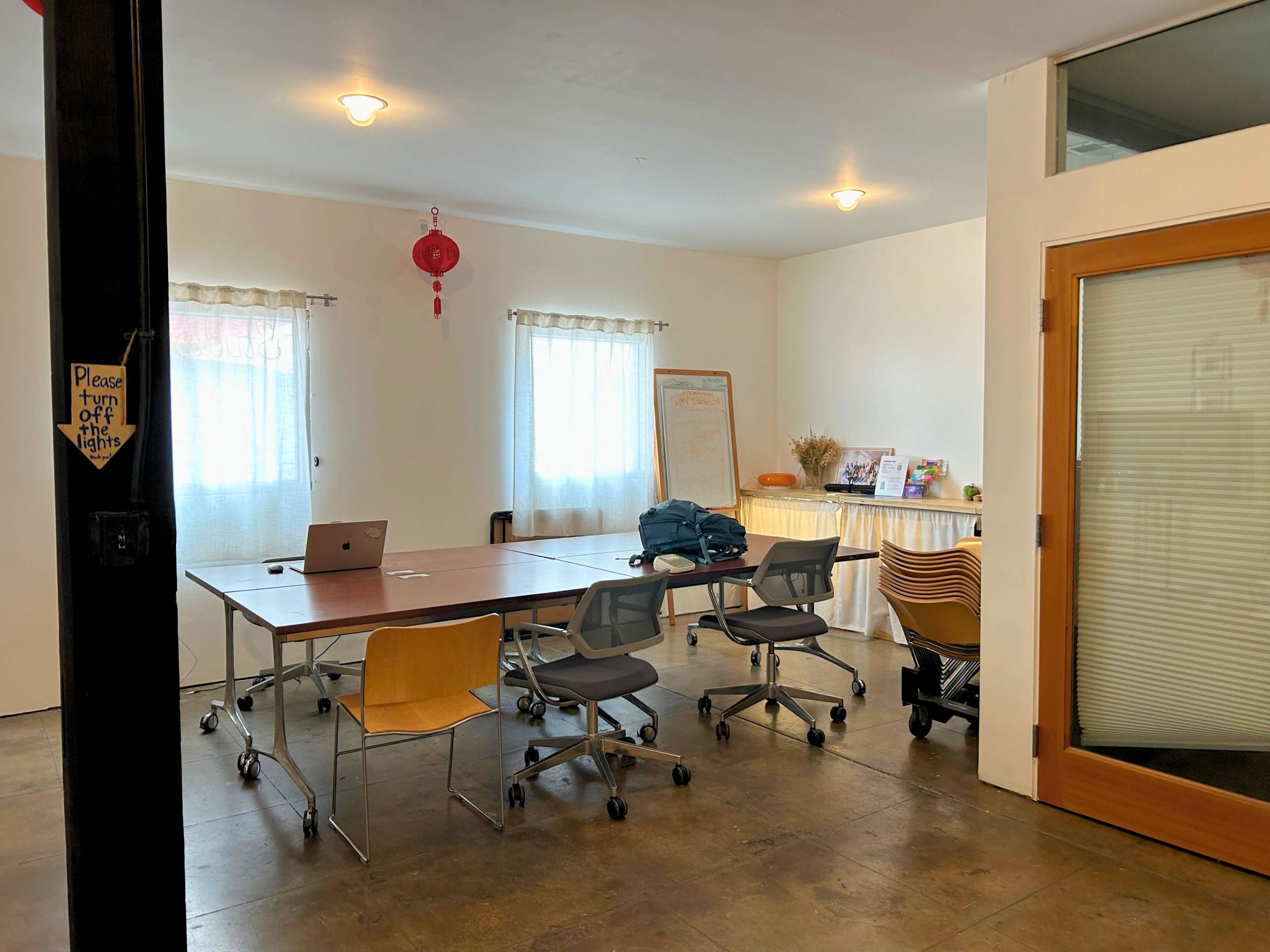 The image shows a light-filled meeting room with a long table surrounded by chairs, a laptop on the table, and windows with sheer curtains.