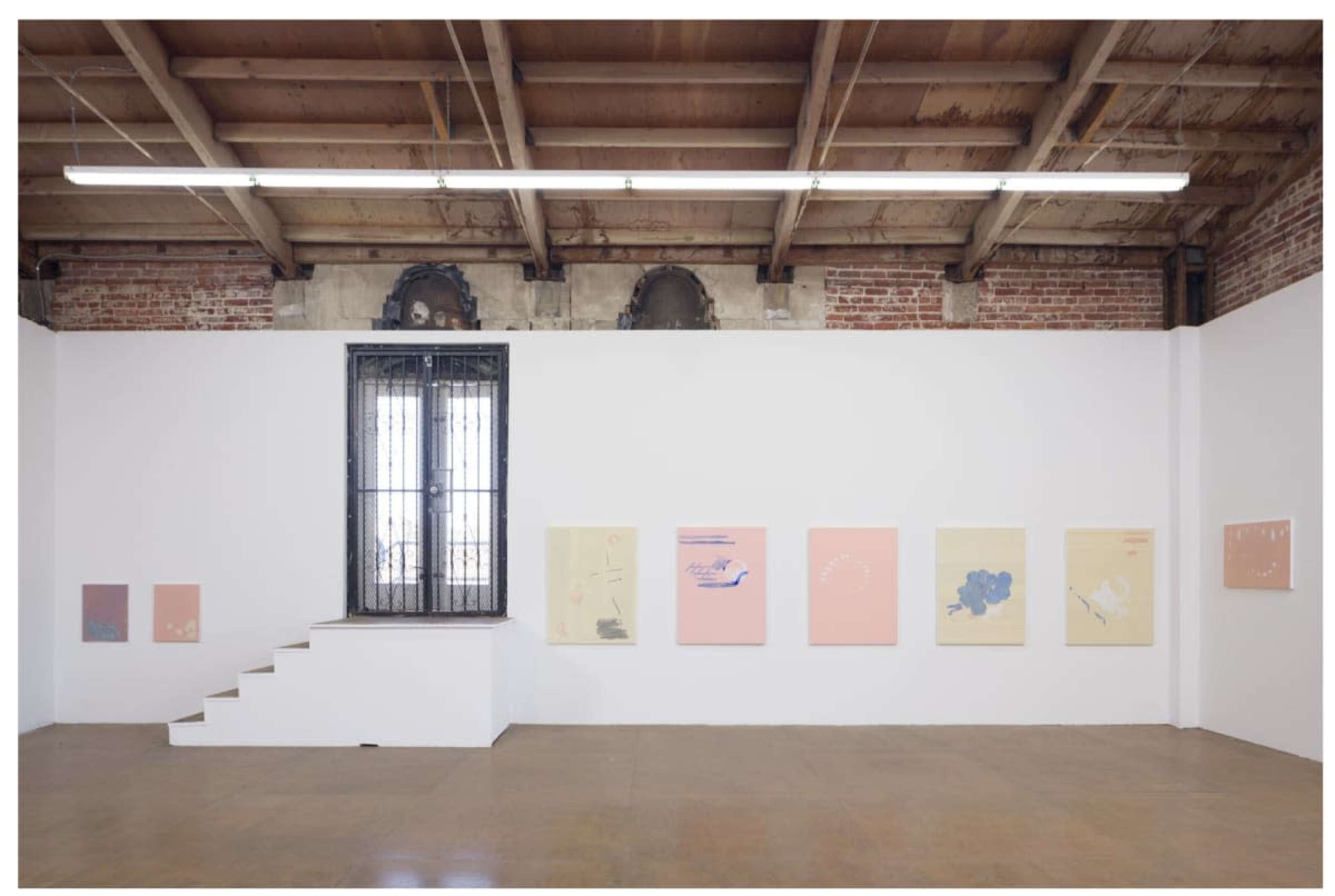 An art gallery with white walls displaying several abstract paintings in soft colors, and a set of stairs leading to a window framed by brick walls.