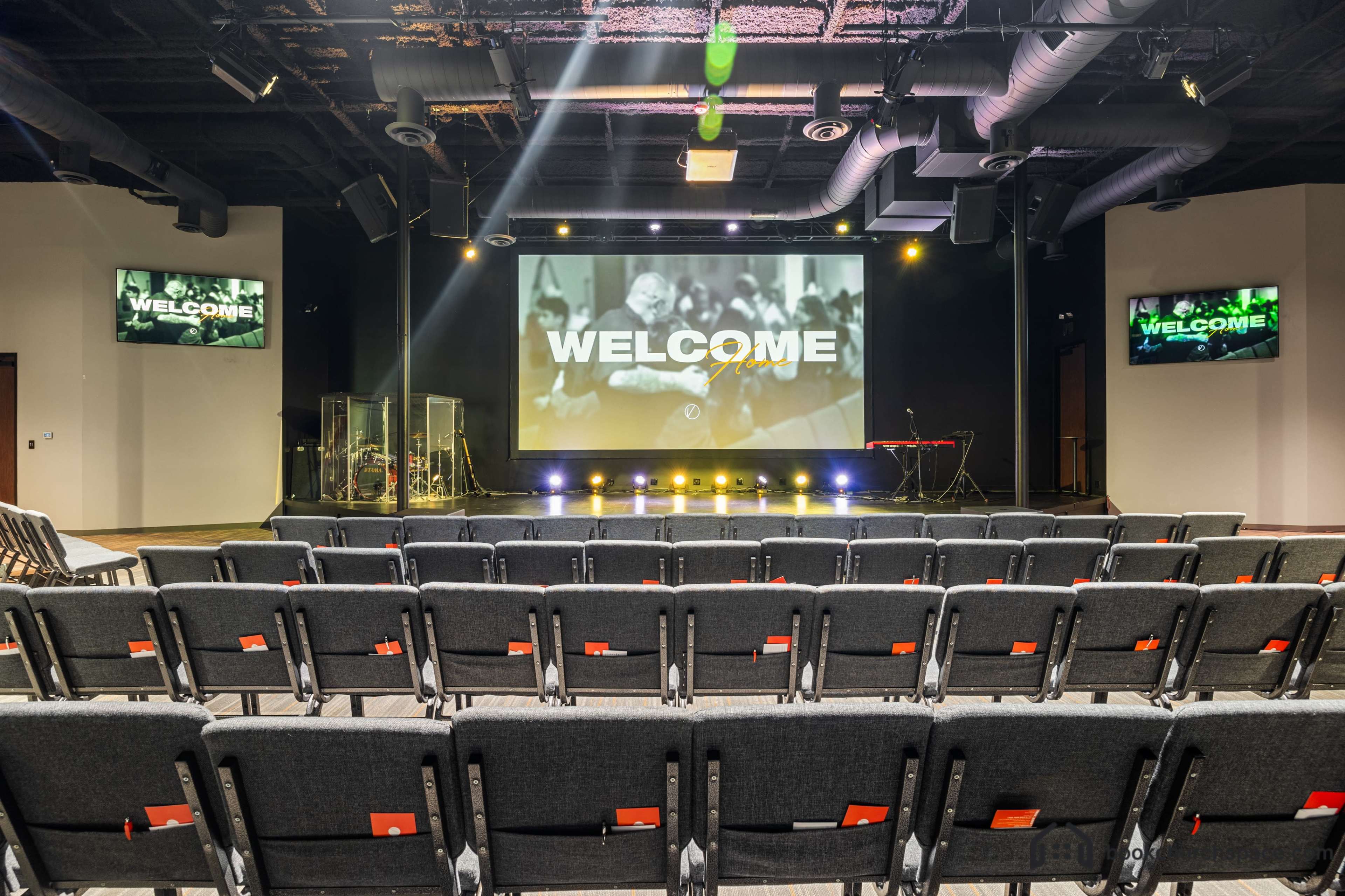 The image shows an auditorium setup with rows of chairs facing a large screen displaying the word "WELCOME" and stage equipment in the background.