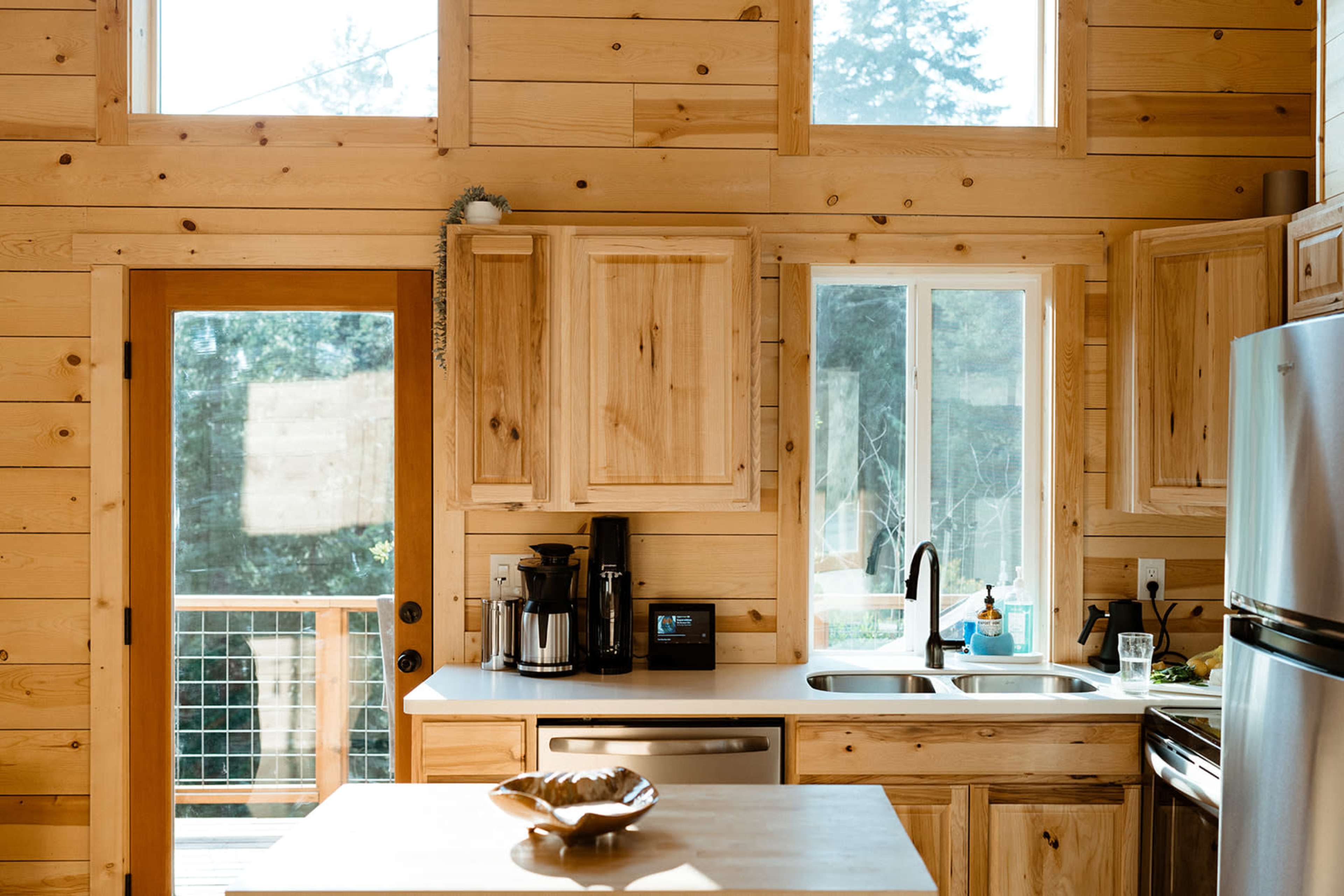 The image shows a wooden kitchen with light-colored cabinets, a countertop, a coffee maker, and a view of a deck through a door and window.