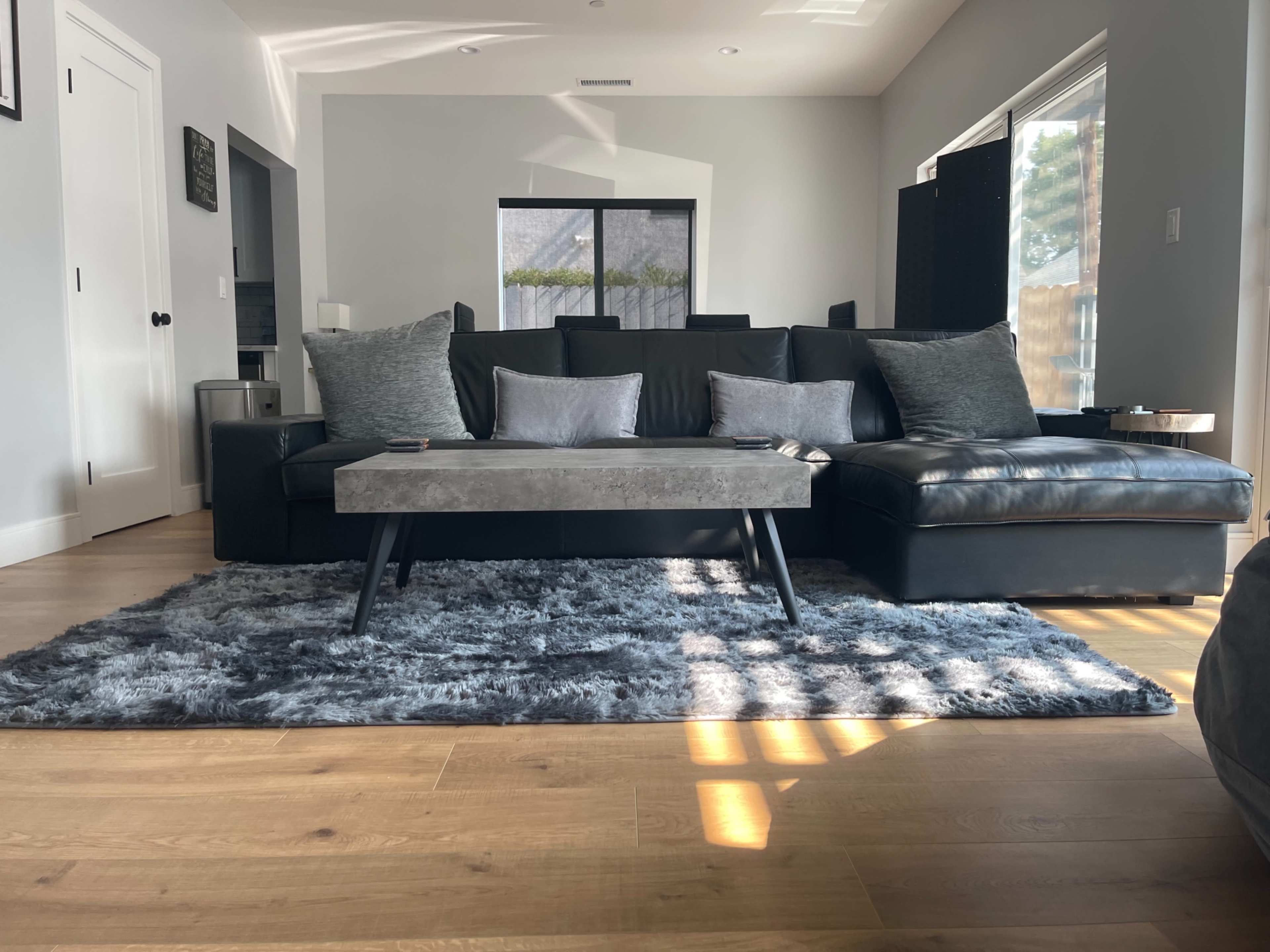 A modern living room with a black sectional sofa, a gray coffee table, and a soft rug on hardwood flooring, illuminated by natural light.