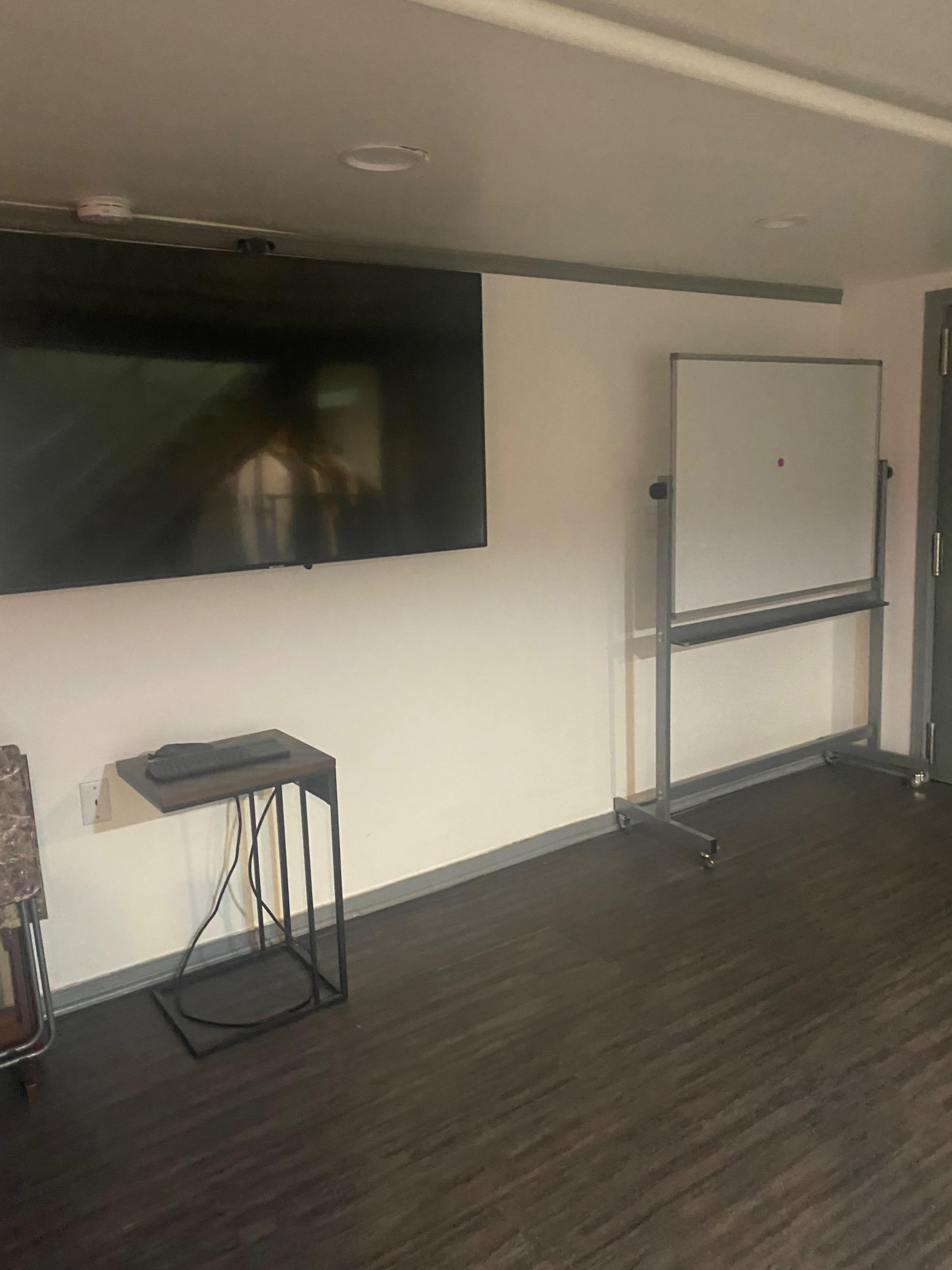 A sparsely furnished room with a flat-screen TV mounted on the wall, a small table beside it, and a movable whiteboard on wheels.