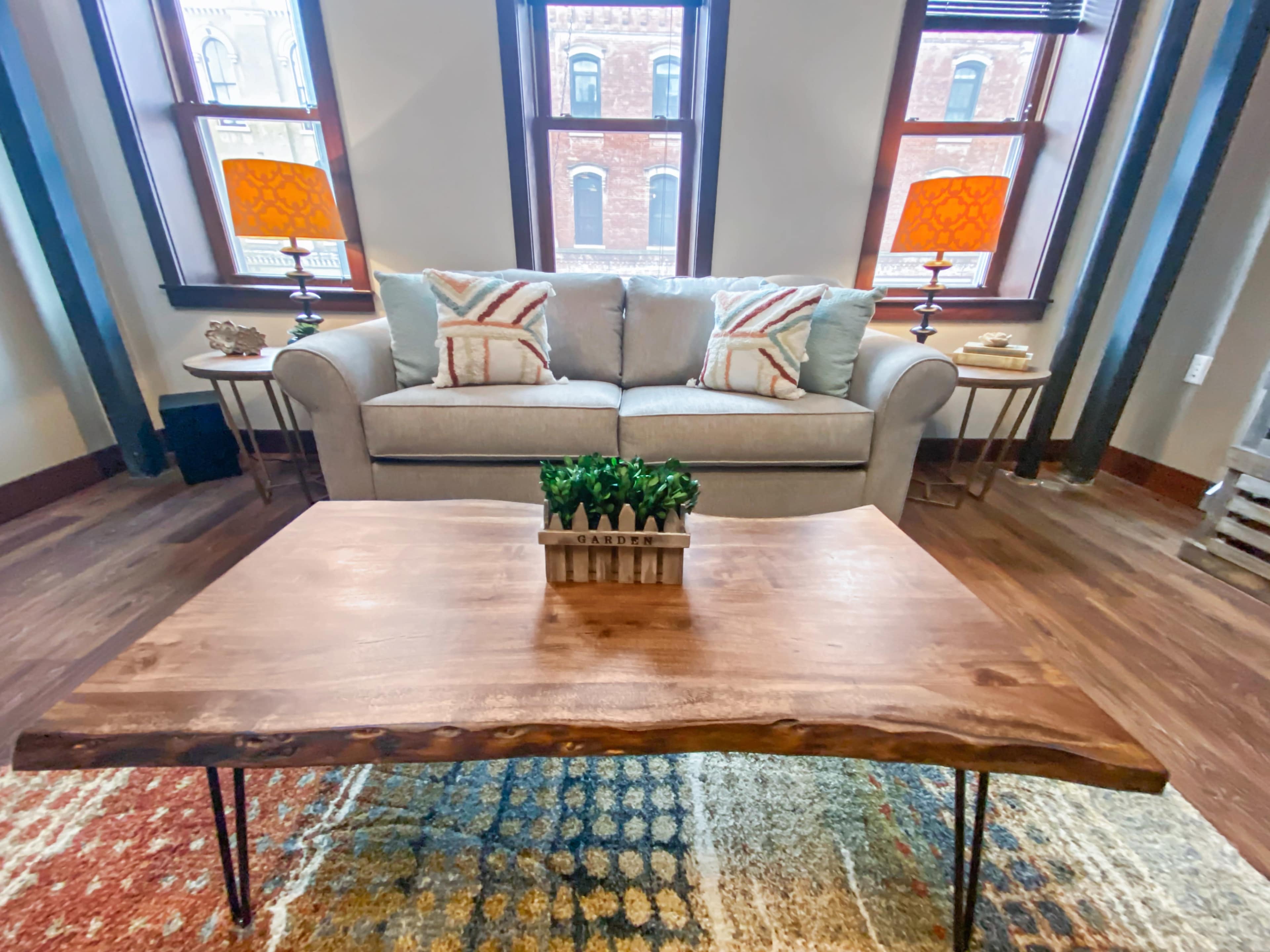 A light gray sofa with decorative pillows is placed in a room featuring a wooden coffee table and two lamps with orange shades beside large windows.