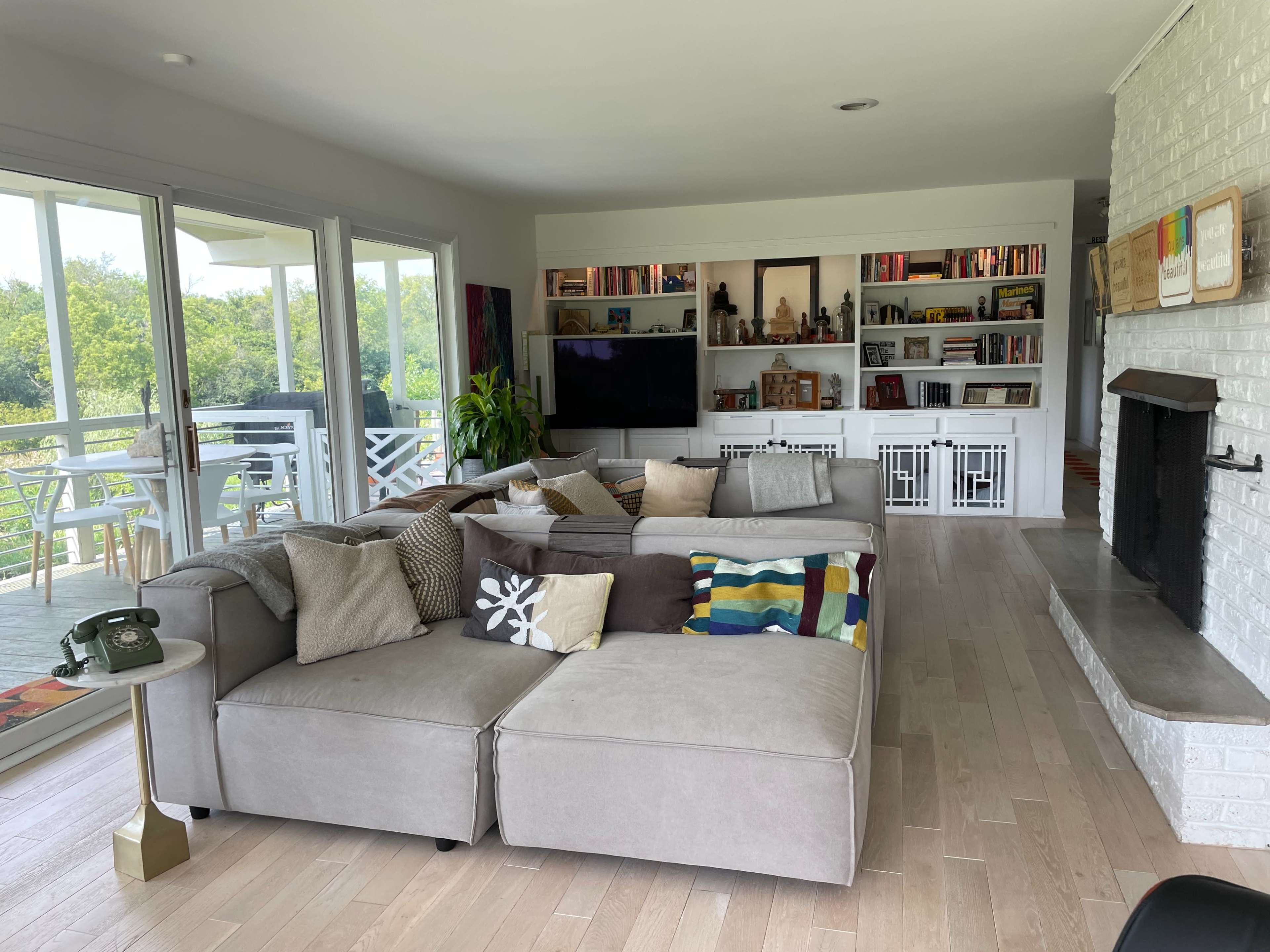 A spacious living room features a large sectional sofa with various pillows, a flat-screen TV, and built-in shelves filled with books and decorative items, alongside a fireplace and large windows opening to an outdoor view.