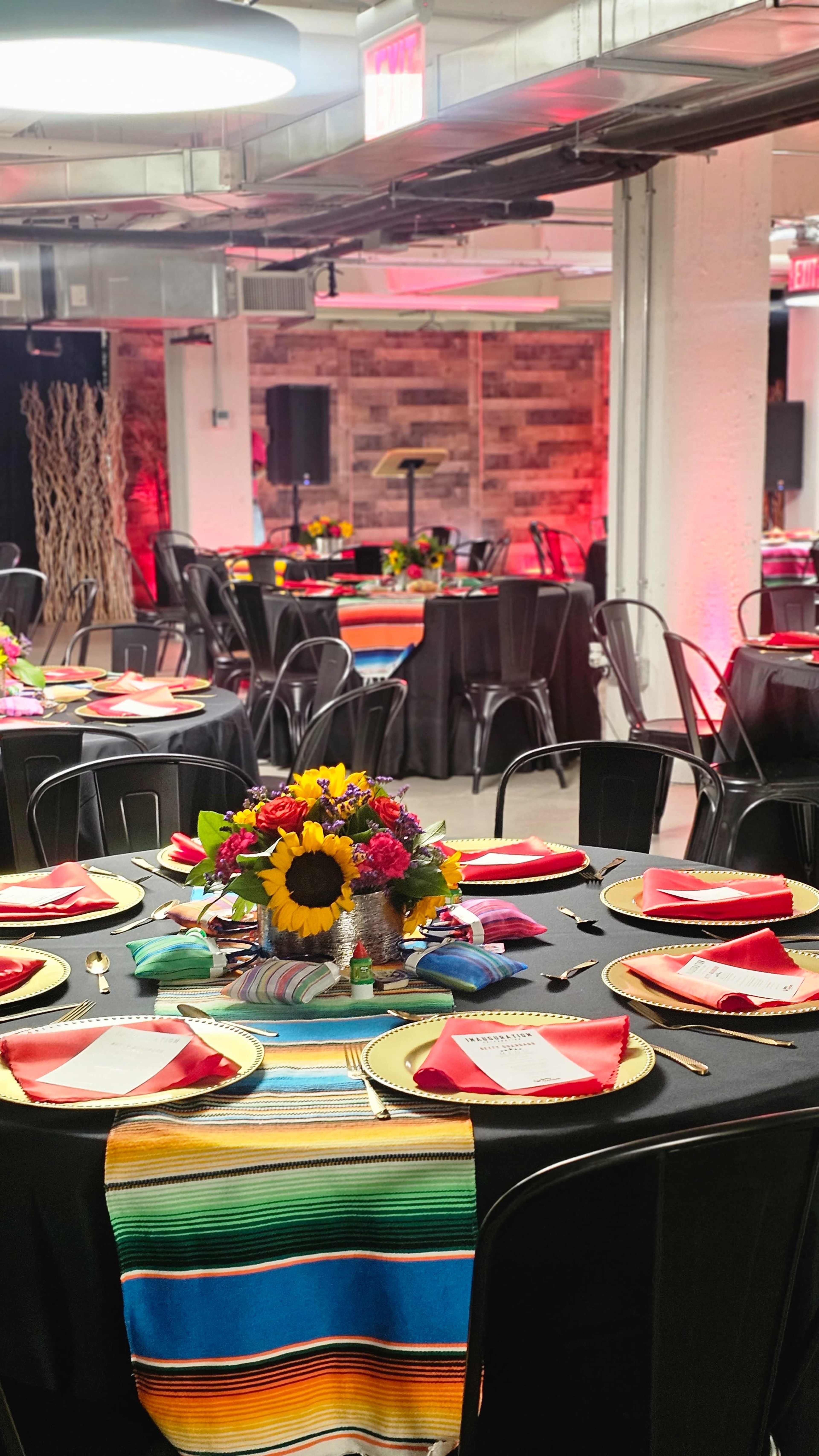 A banquet hall is set up with multiple round tables adorned with colorful tablecloths, gold chargers, and floral centerpieces, illuminated by soft red lighting.