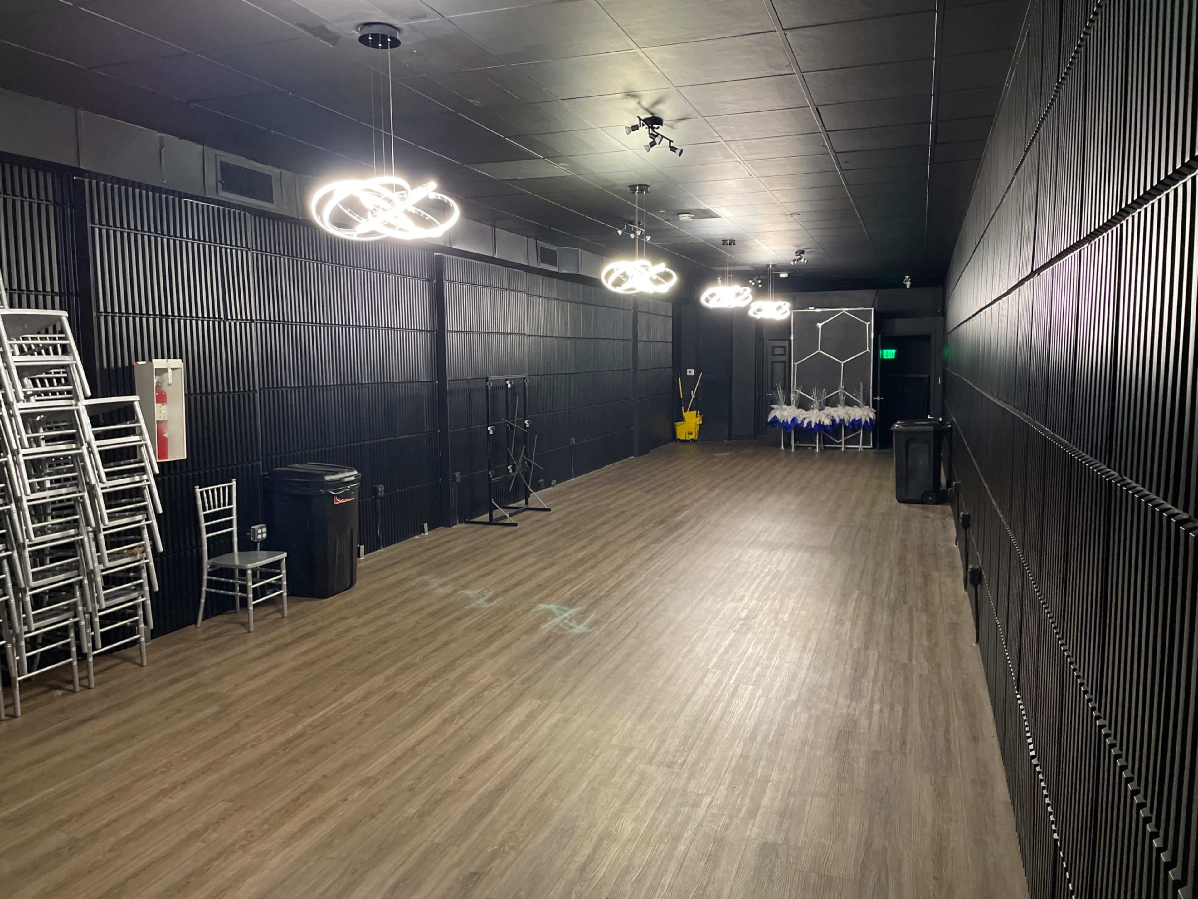 An empty, elongated room with wooden flooring, black panelled walls, and several overhead light fixtures.