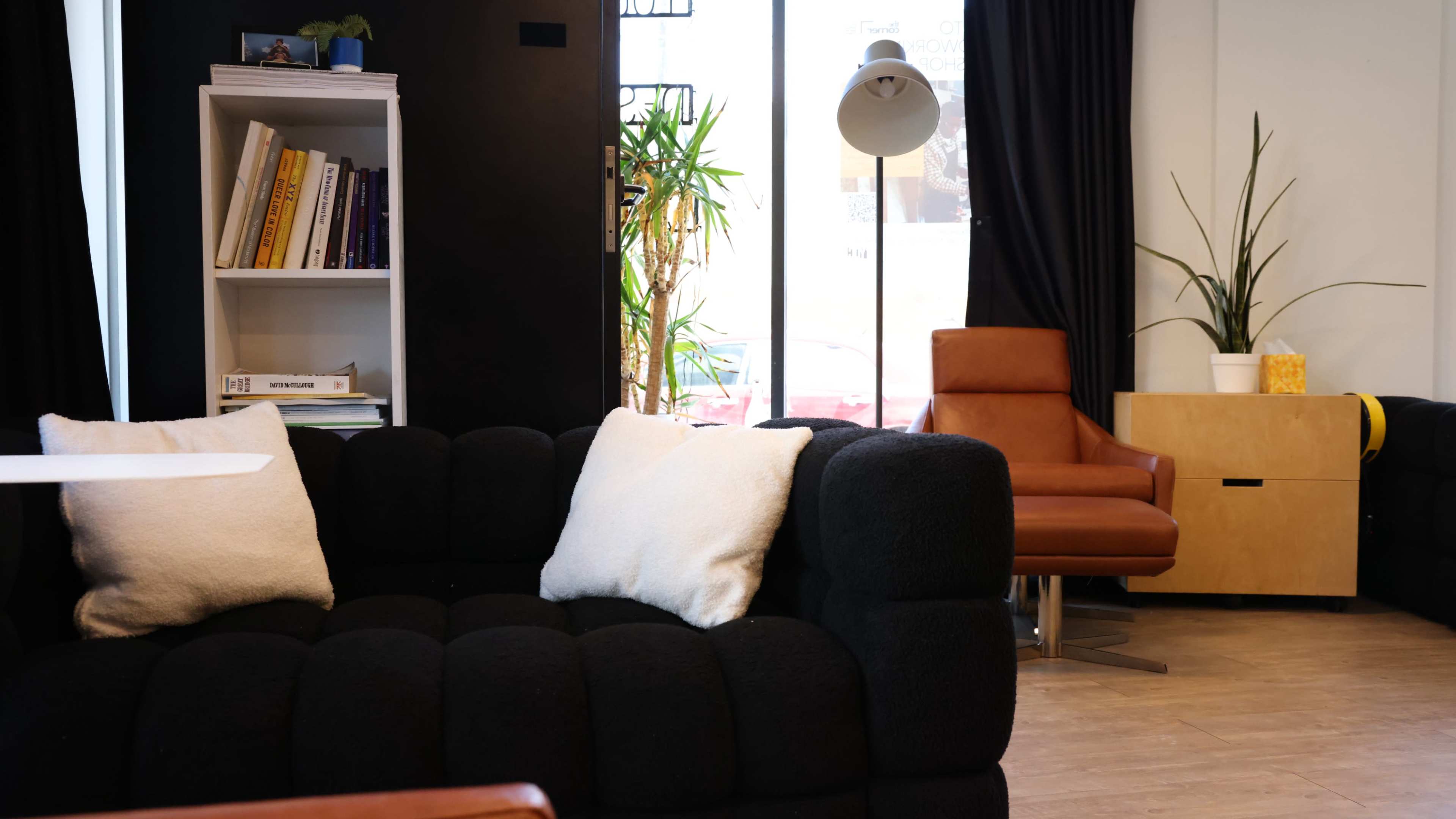 The image shows a cozy interior space with a black upholstered sofa, a brown leather chair, a white bookshelf filled with books, and large windows allowing natural light to enter.