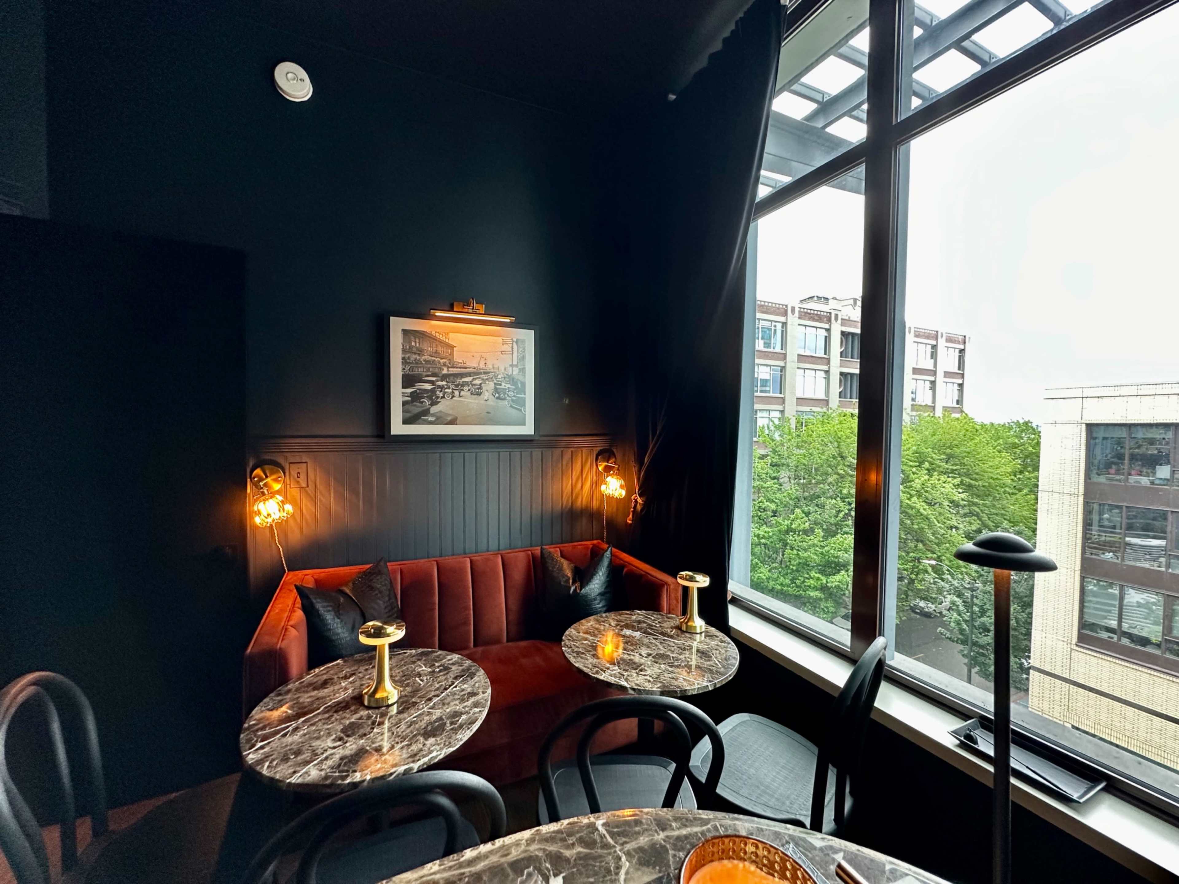 A cozy seating area features a plush red couch and marble-topped tables beside a large window overlooking a tree-lined street.