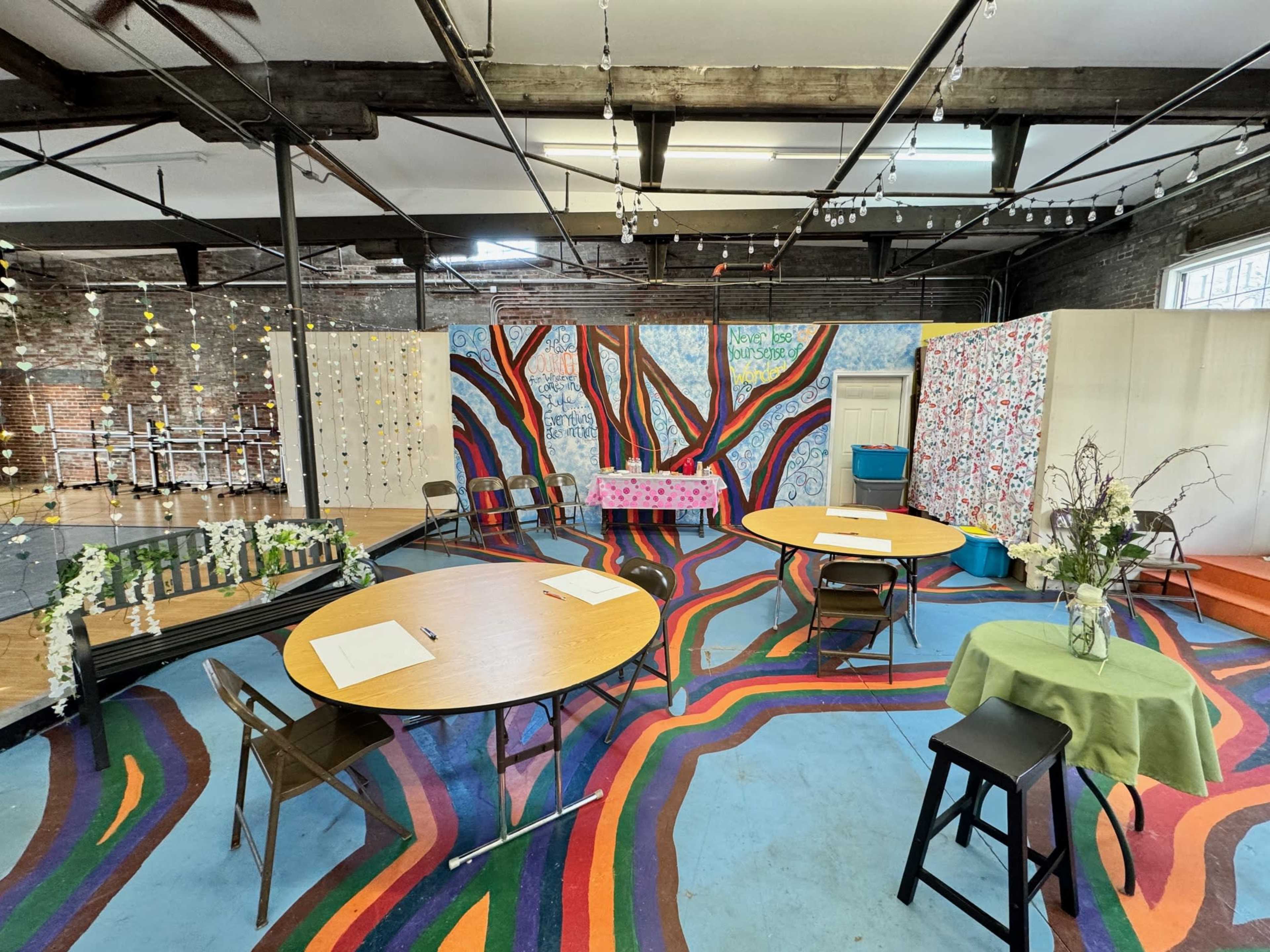 A colorful, multi-purpose room with patterned flooring, several circular tables, chairs, and decorated walls.