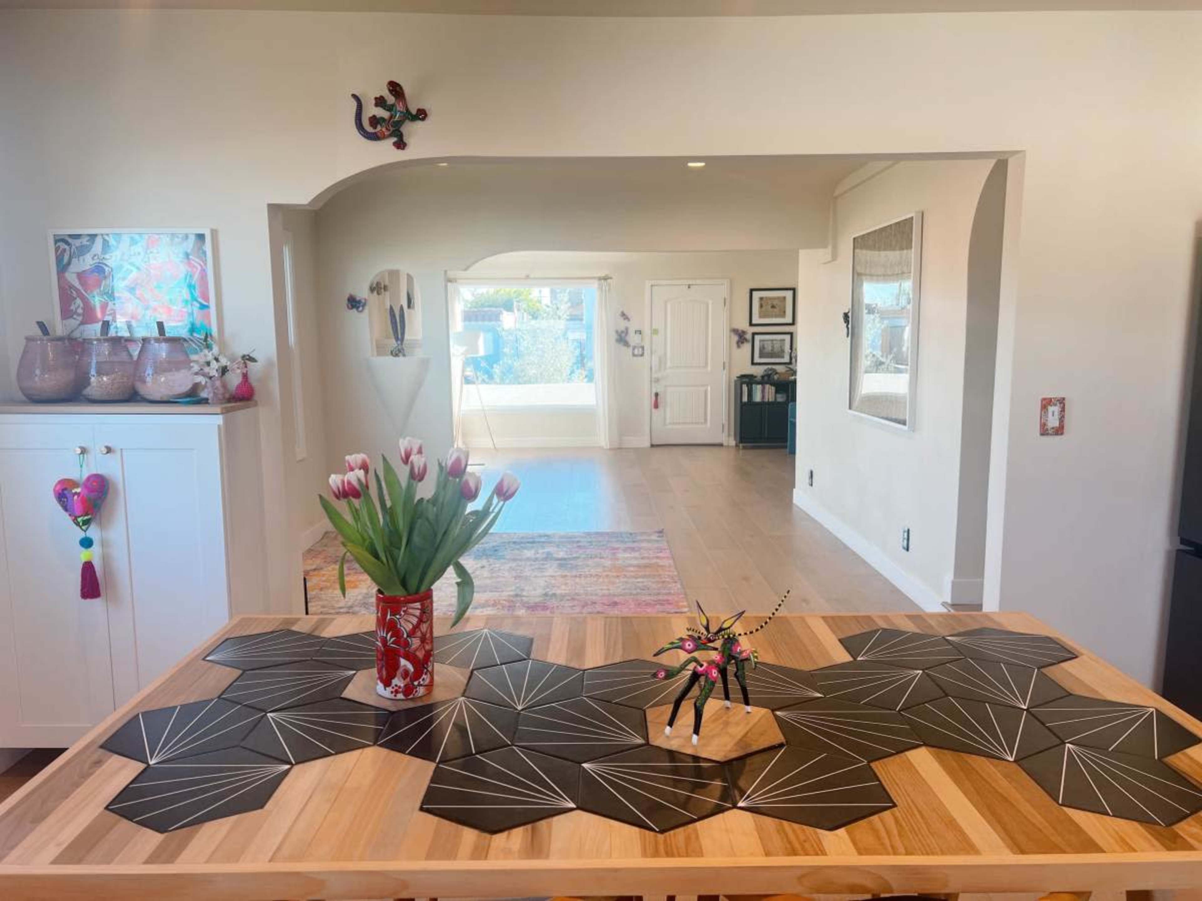 A wooden table with a geometric black and gray placemat holds a vase of tulips, while a doorway leads into a well-lit living space featuring a colorful rug.