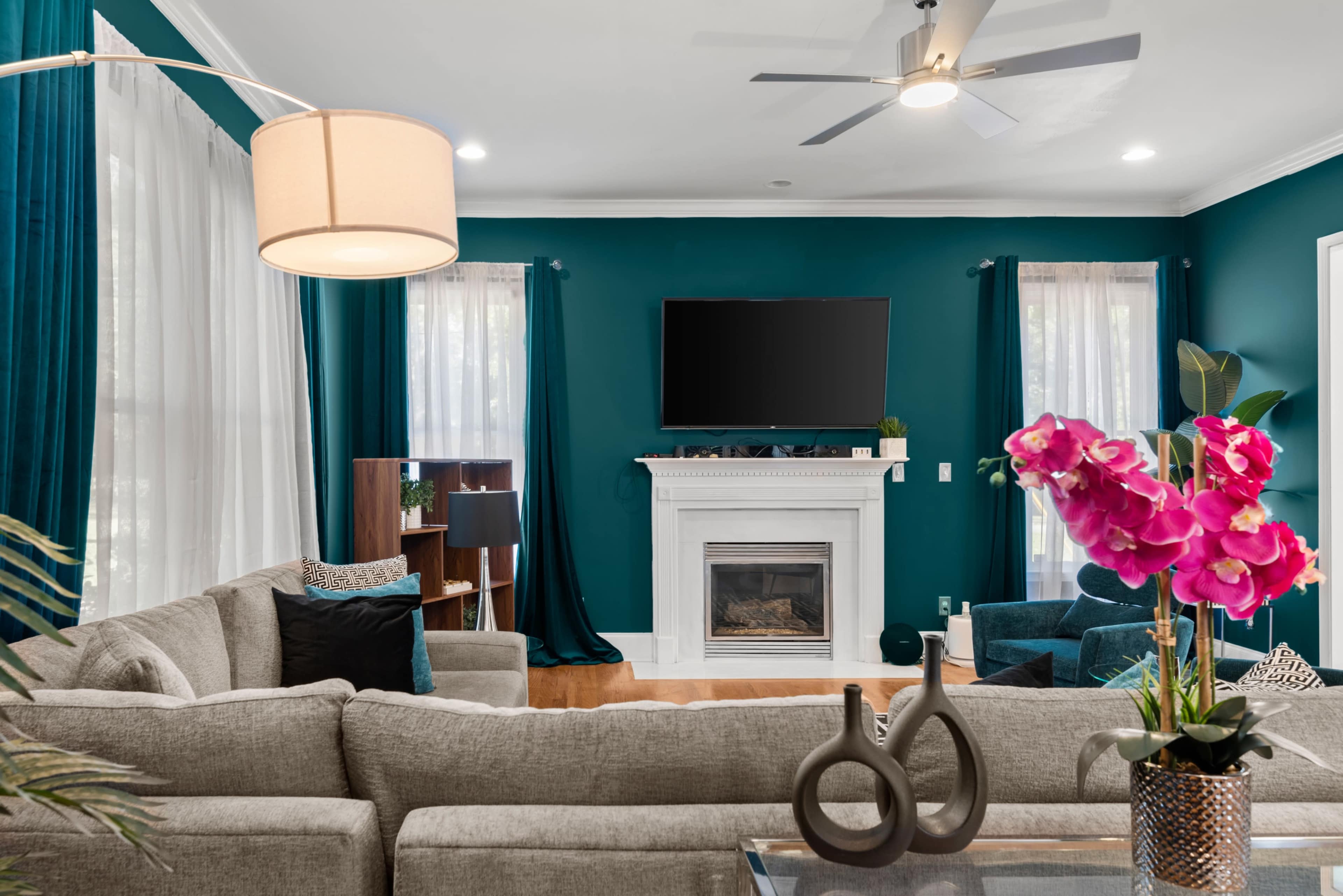 A modern living room features a grey sectional sofa, a fireplace with a TV above it, and teal curtains framing the windows.