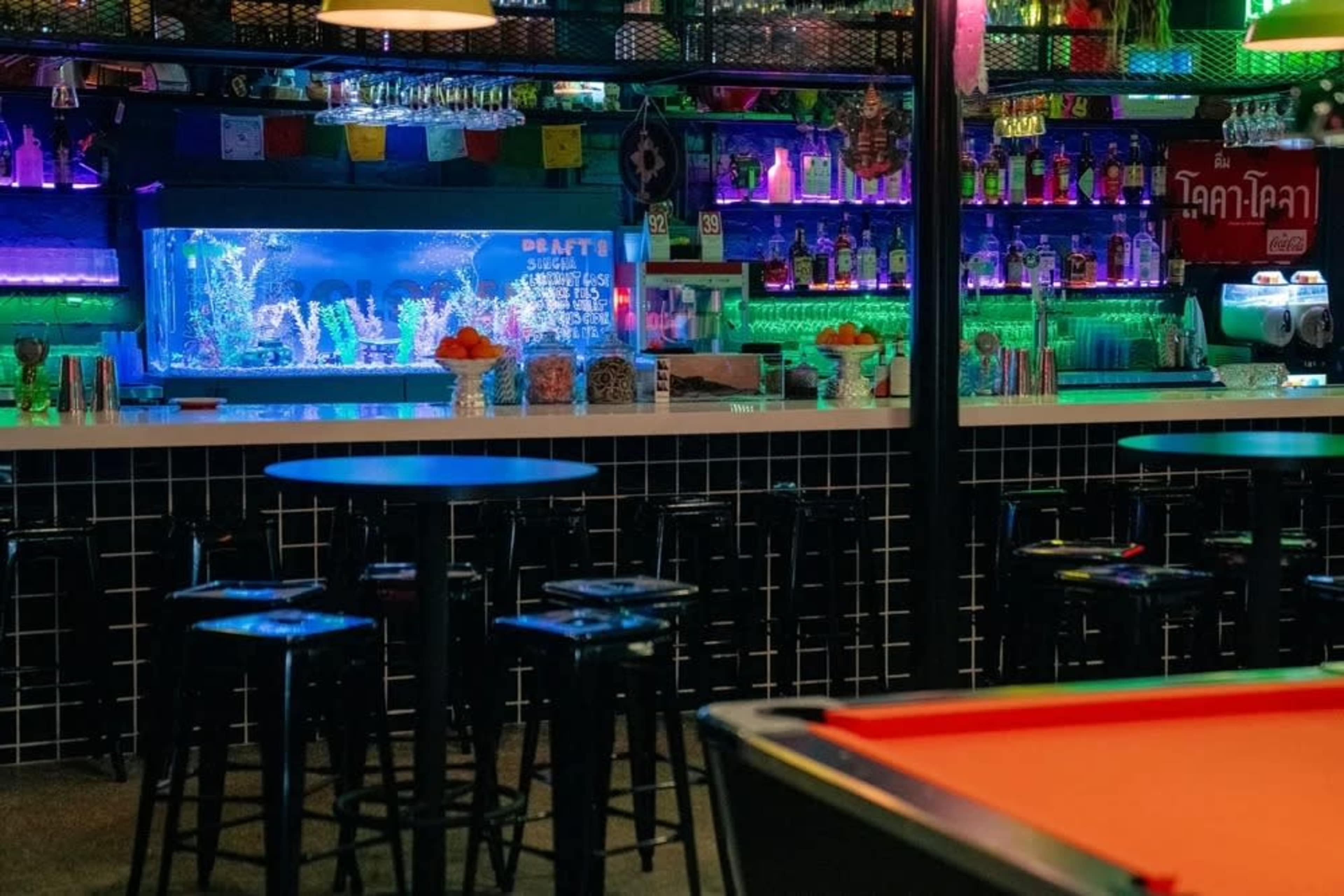 A brightly lit bar features colorful neon lights and a fish tank behind the counter, with bar stools arranged in front of a pool table.
