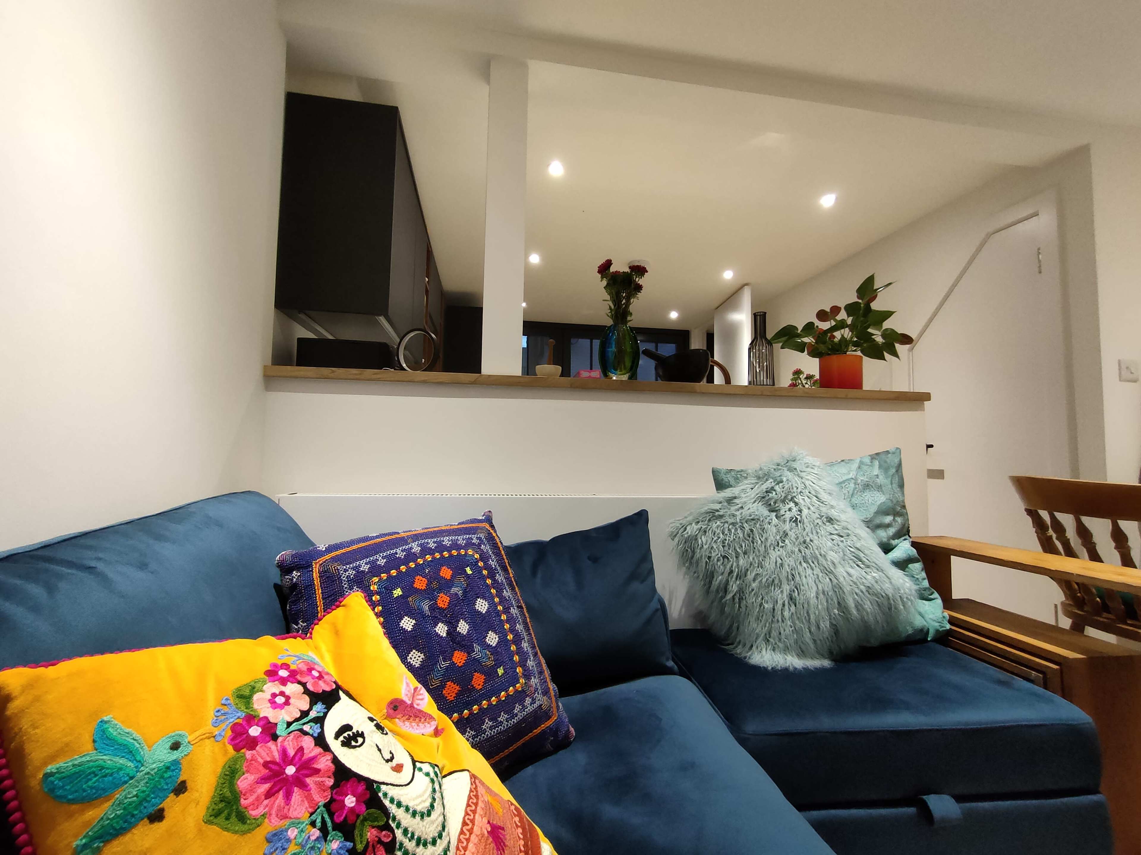 A blue sofa with colorful cushions is positioned in a well-lit living room, featuring a kitchen area and decorative plants in the background.