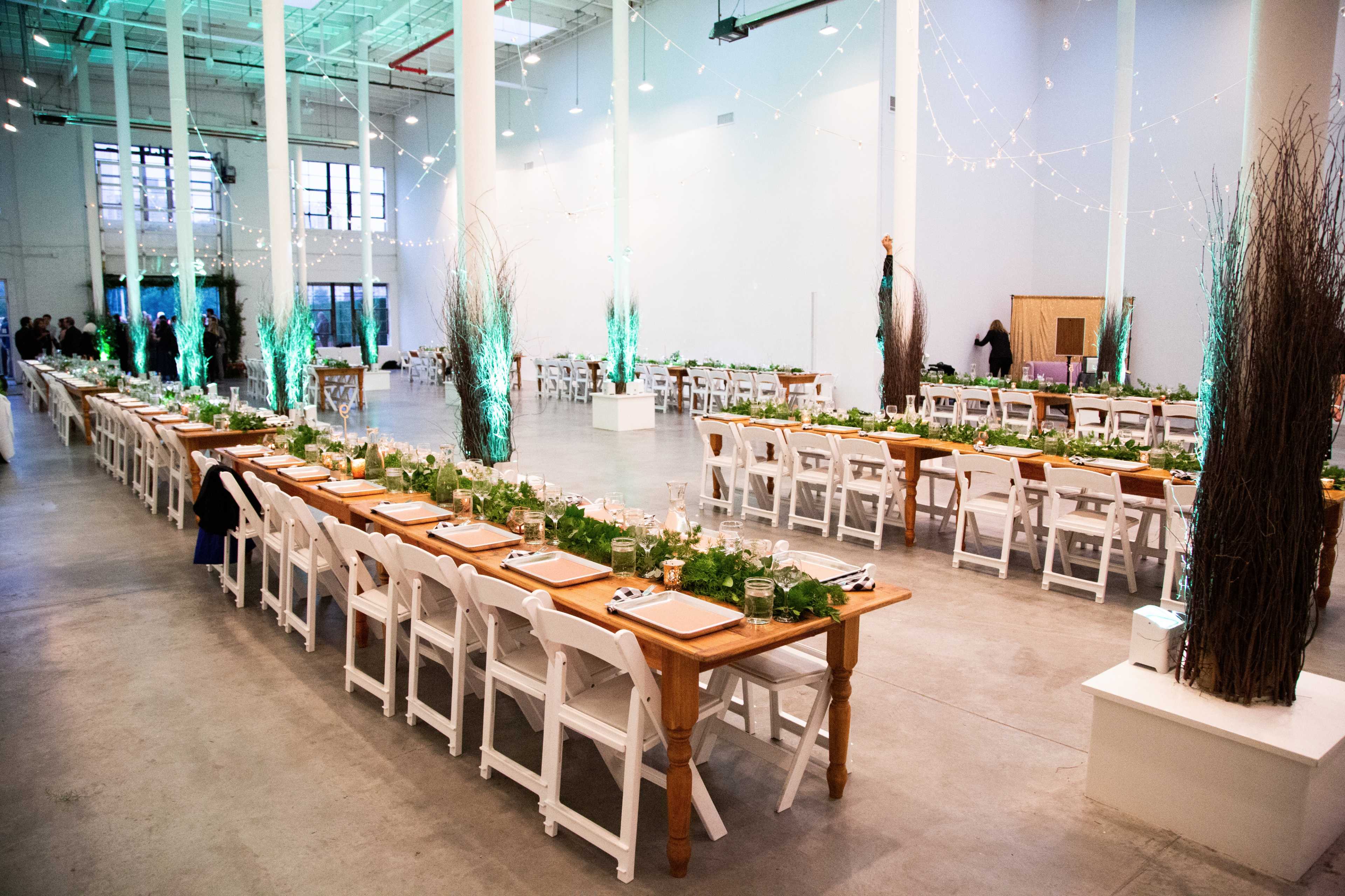A spacious indoor venue features long tables set for a dinner event, adorned with greenery and illuminated by string lights.