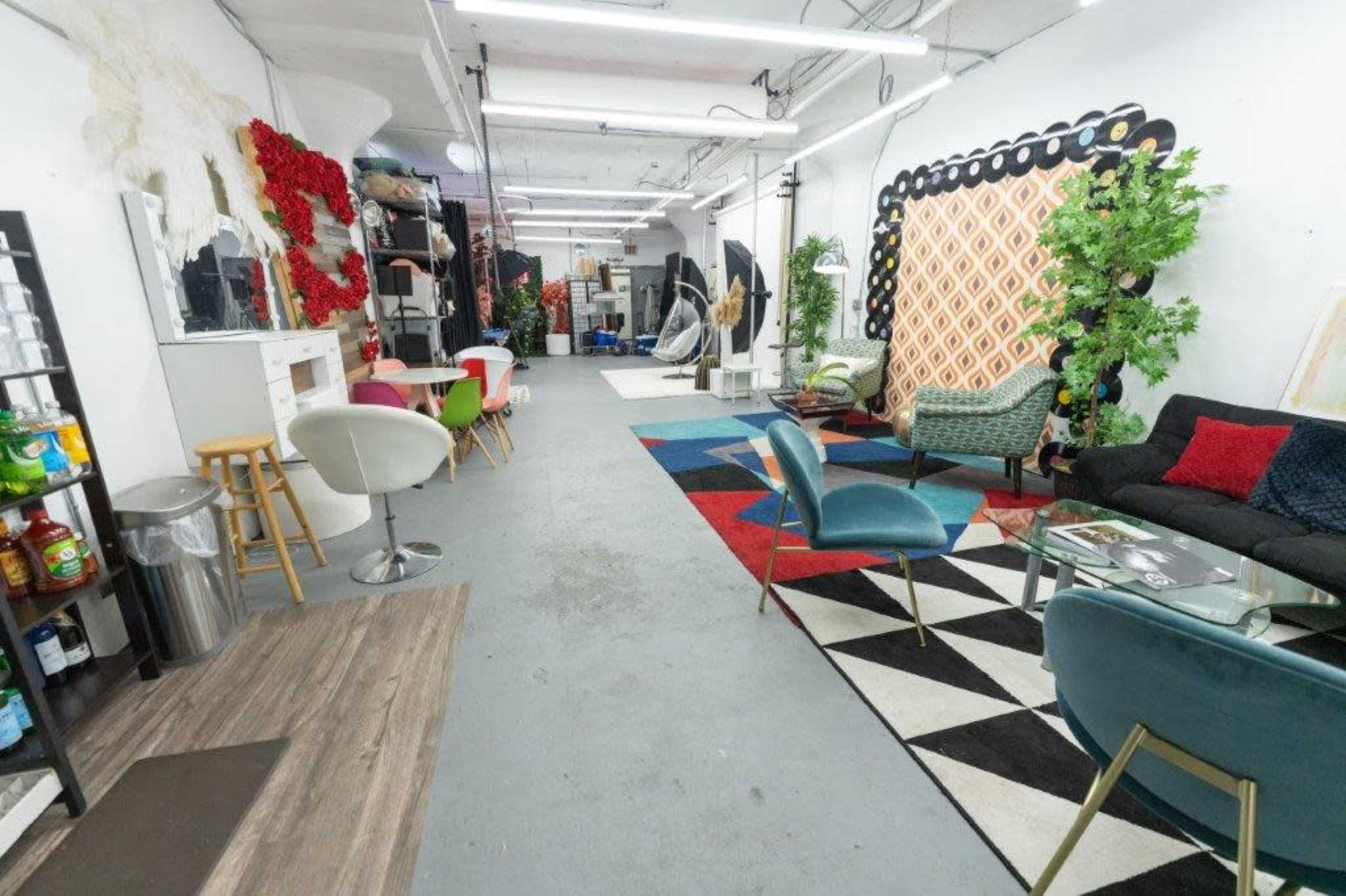 A spacious studio interior with various seating areas, decorative plants, and colorful rugs, along with equipment like lights and backdrops.