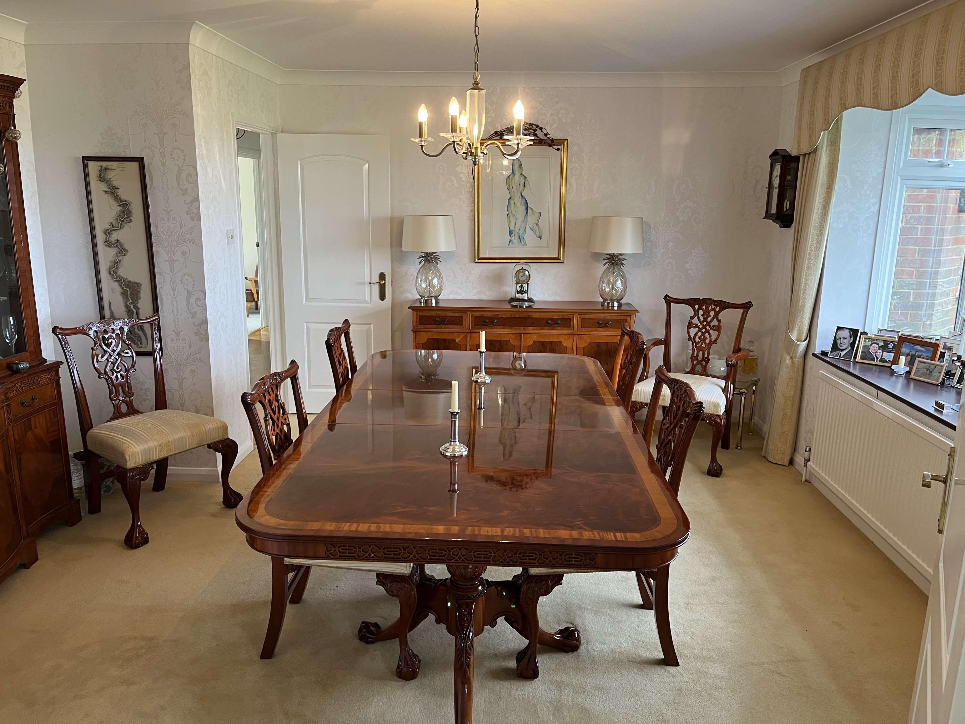 An elegantly arranged dining room featuring a large wooden table surrounded by six chairs, with a chandelier overhead and decorative lamps on a sideboard.