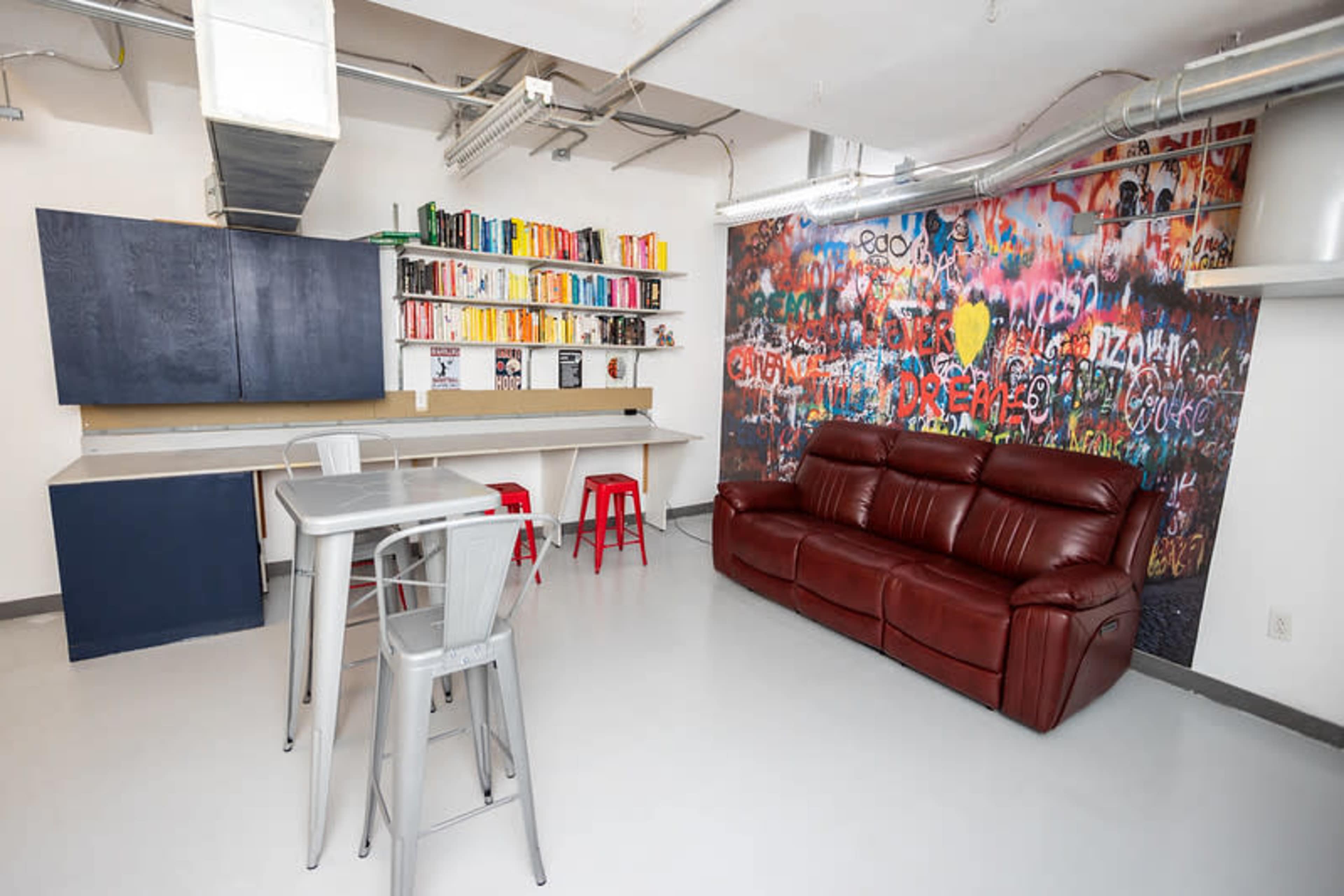 The image shows a modern, minimalist room featuring a kitchen area with blue cabinetry, a colorful bookshelf, a grey dining table with metal chairs, and a brown leather couch against a vibrant graffiti wall.