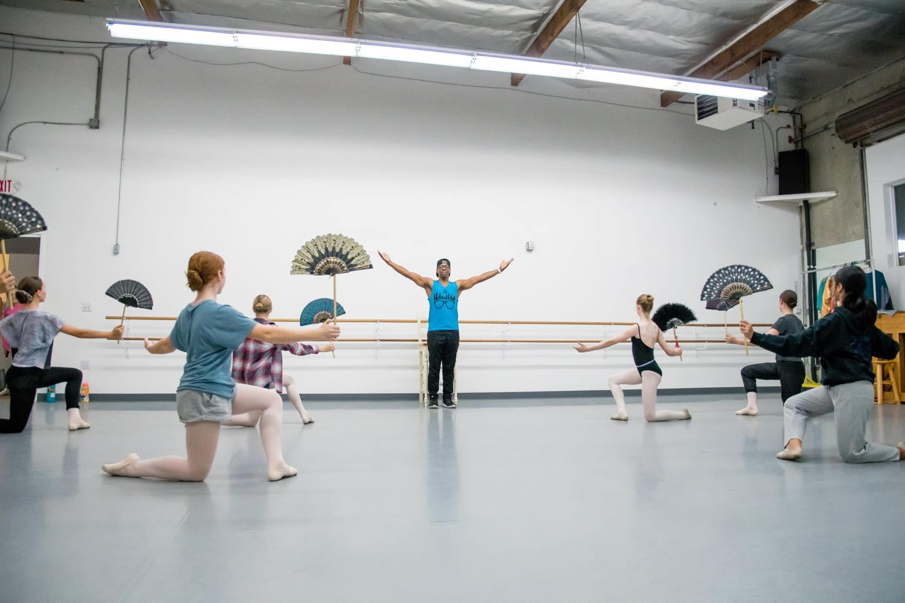 A group of dancers practices in a studio while holding fans, with an instructor positioned in front, raising his arms.