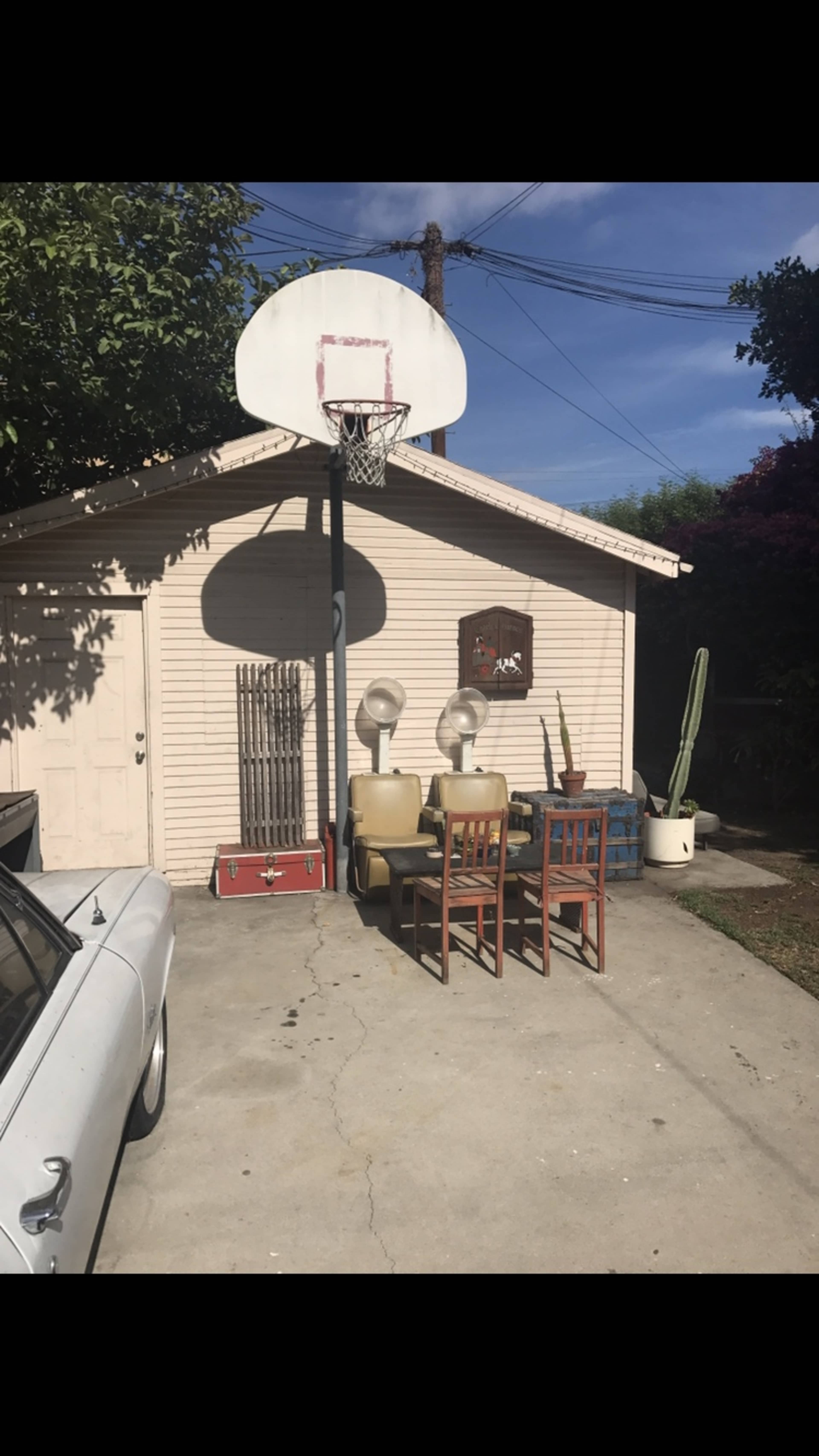 A backyard scene with a basketball hoop attached to a garage, a set of chairs and a table nearby, and several plants around the area.