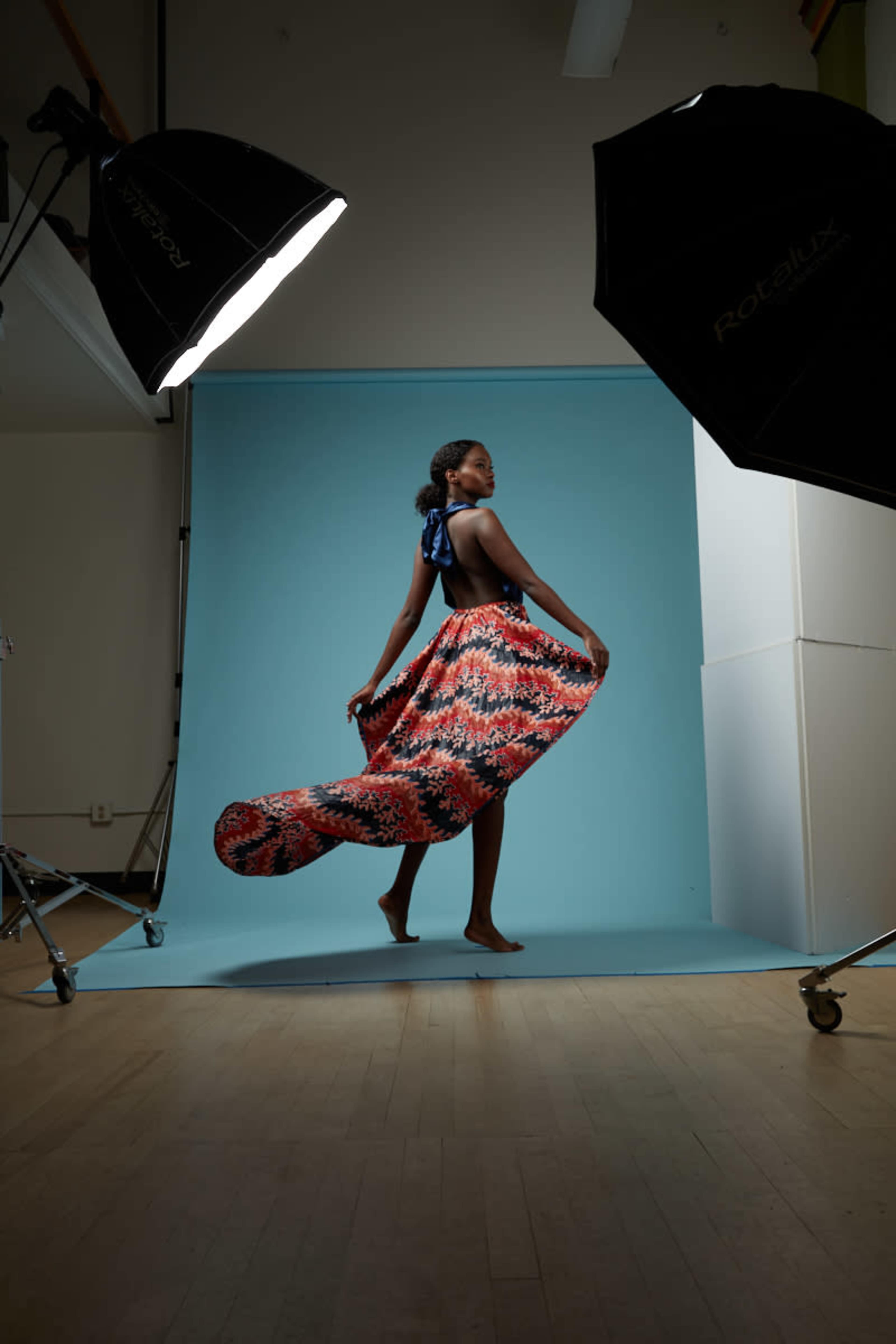 A model poses gracefully in a colorful flowing dress against a light blue backdrop, surrounded by studio lighting equipment.