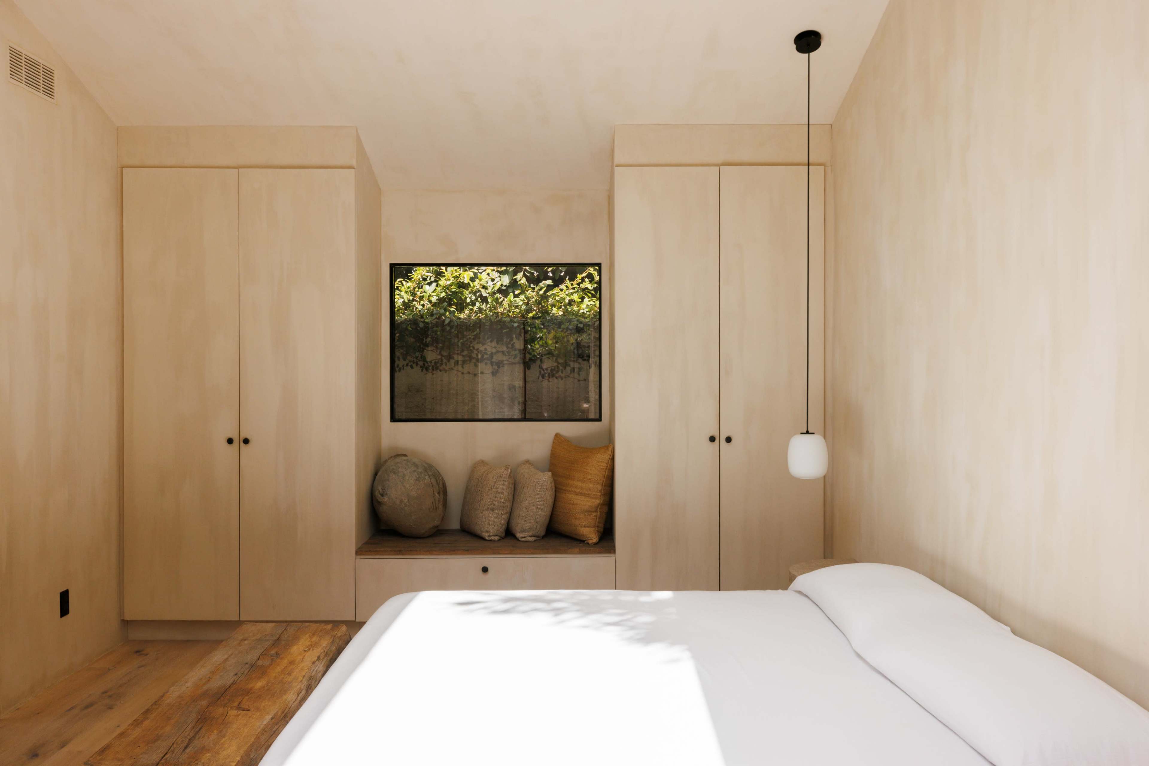 A minimalist bedroom with wooden floors, built-in cabinetry, and a window seat featuring decorative pillows.
