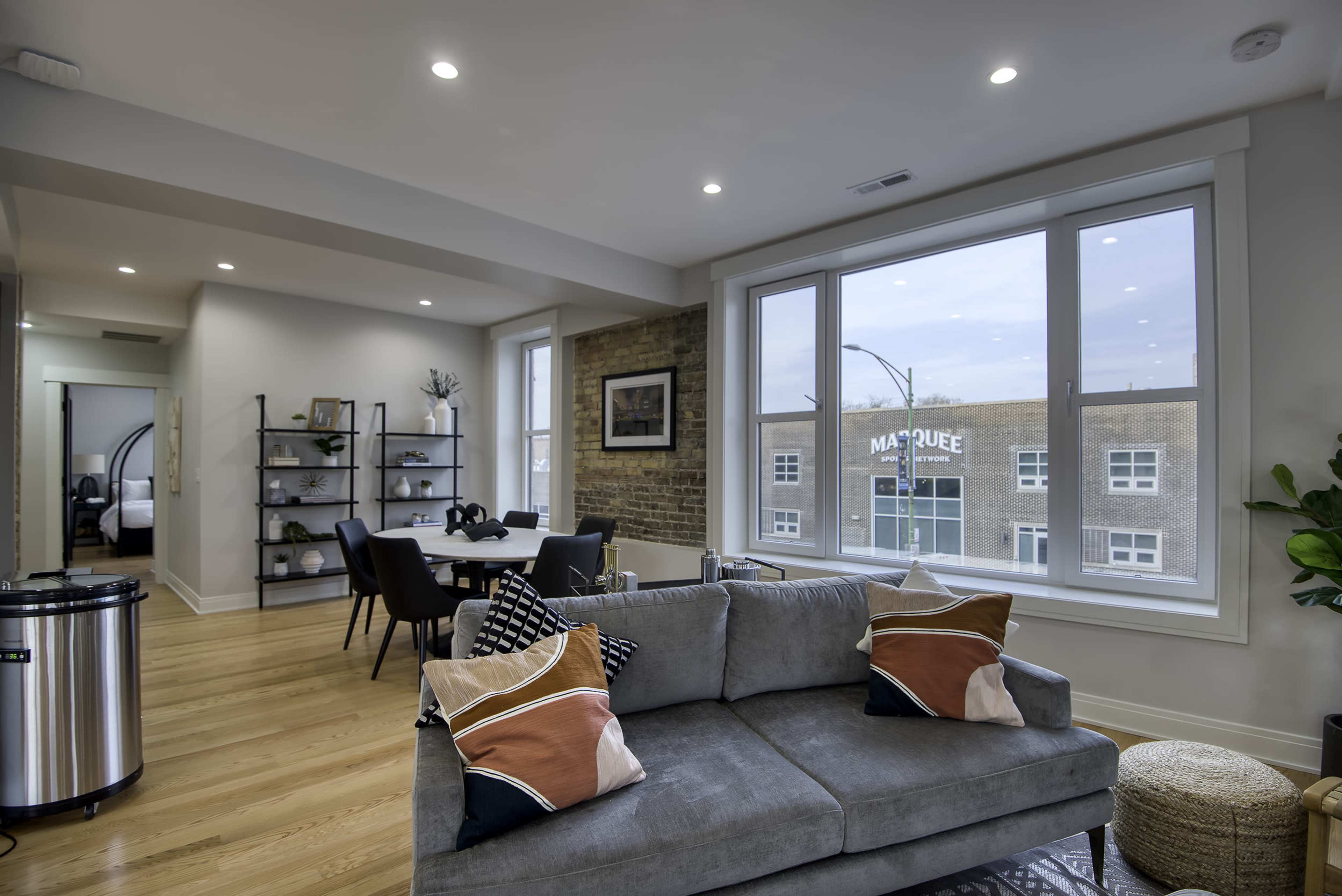 A modern living room features a gray sofa with decorative pillows, a wooden dining table, and large windows overlooking a street with a building named "Marquee."