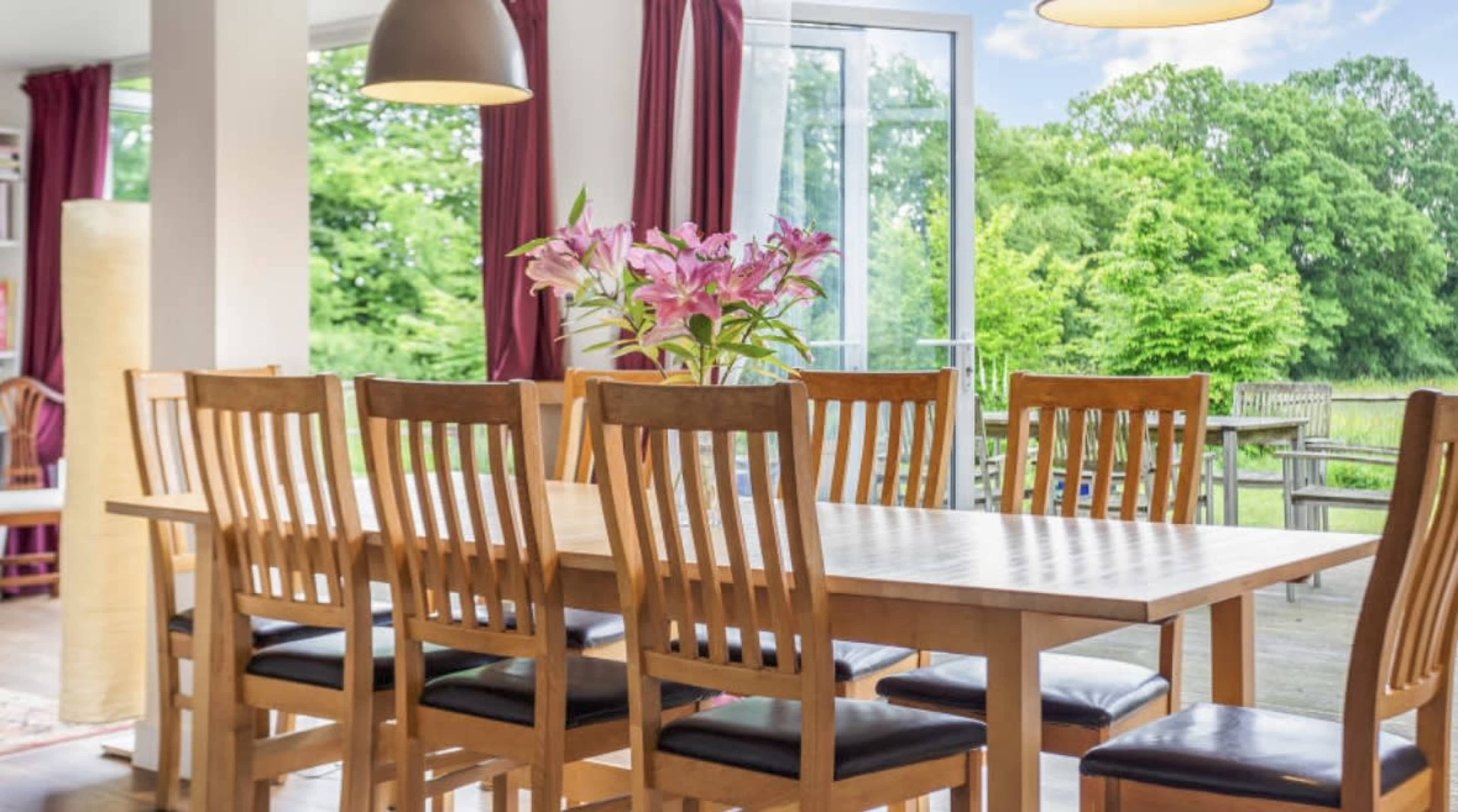 A wooden dining table with eight chairs is set in a brightly lit room, featuring large windows that overlook a green outdoor landscape.
