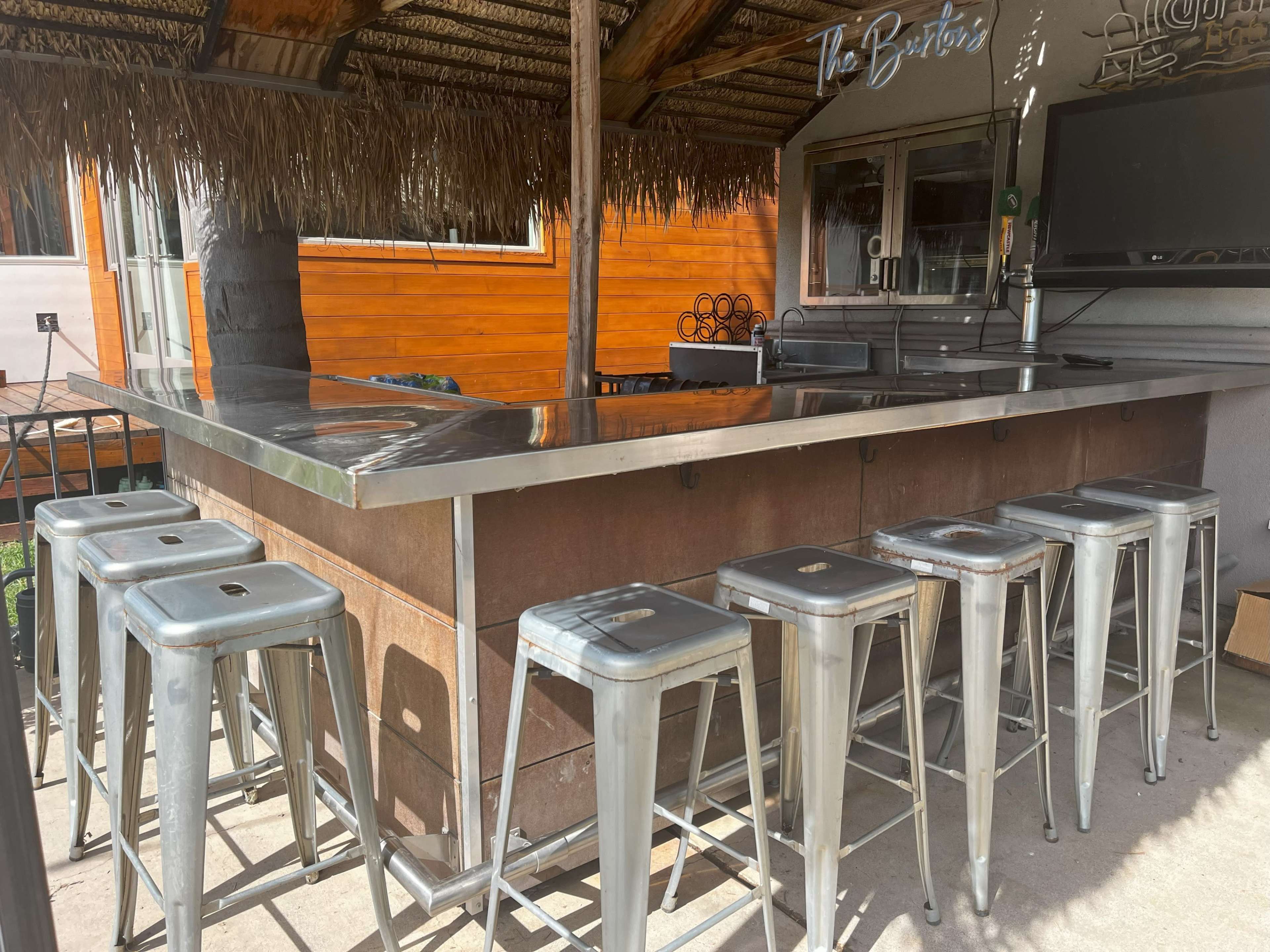 An outdoor bar with a metal countertop and several metal stools aligned in front.