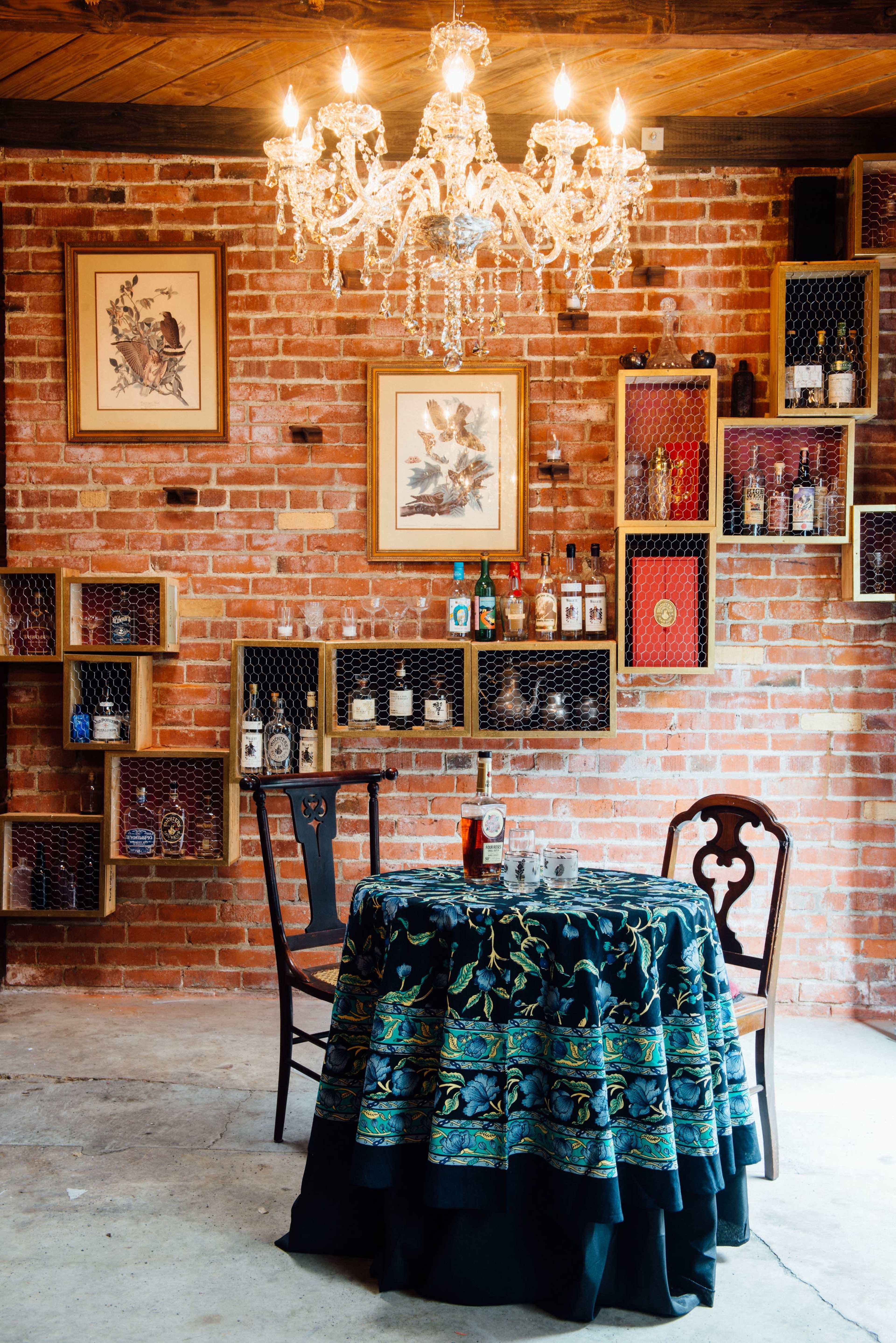 A table adorned with an embroidered tablecloth sits under a chandelier in a brick-walled room featuring shelves of bottles and framed artwork.