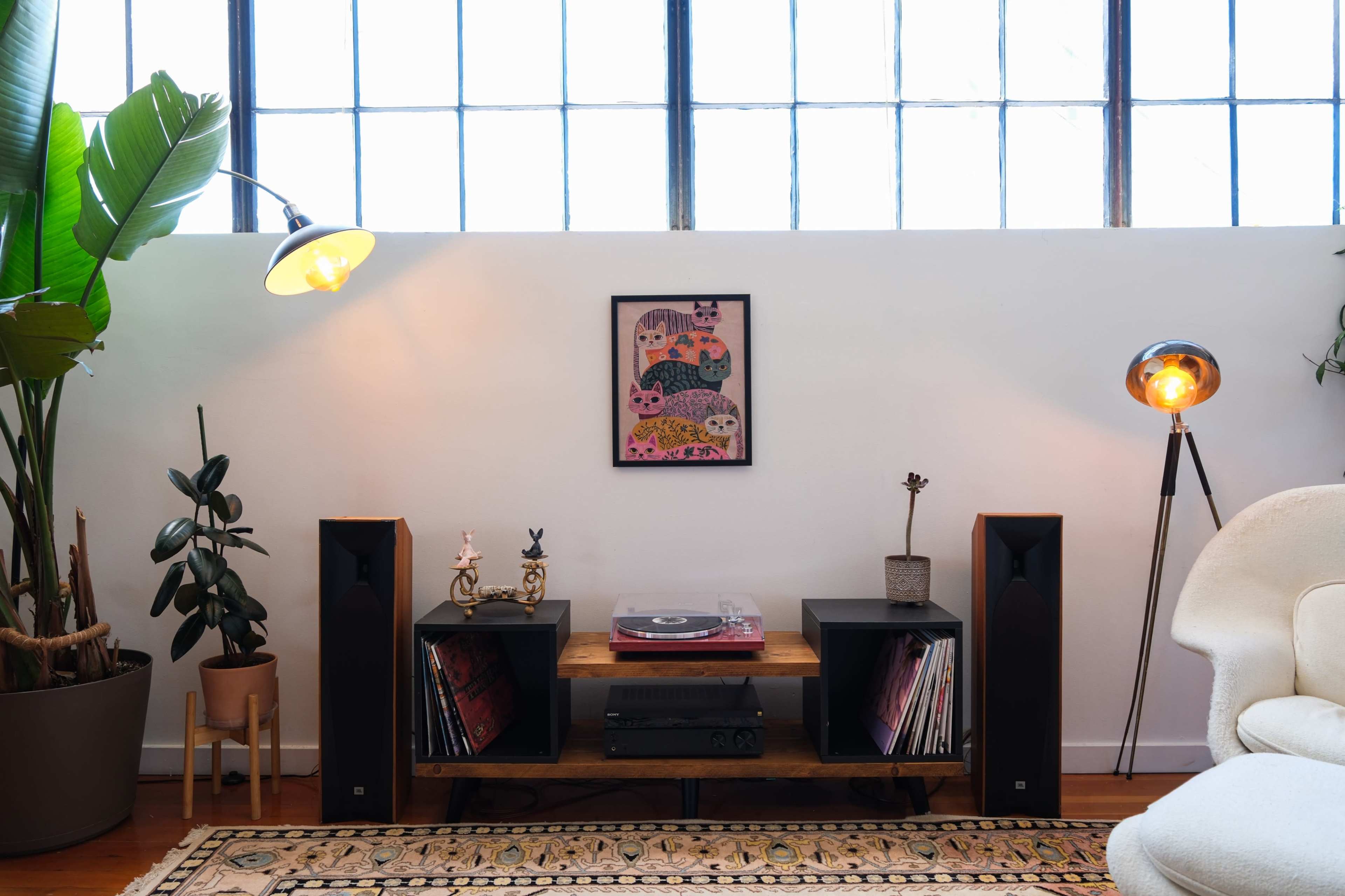 A modern living room features a record player and vinyl collection on a wooden shelf, flanked by tall speakers and plants, beneath a colorful framed artwork and large windows.