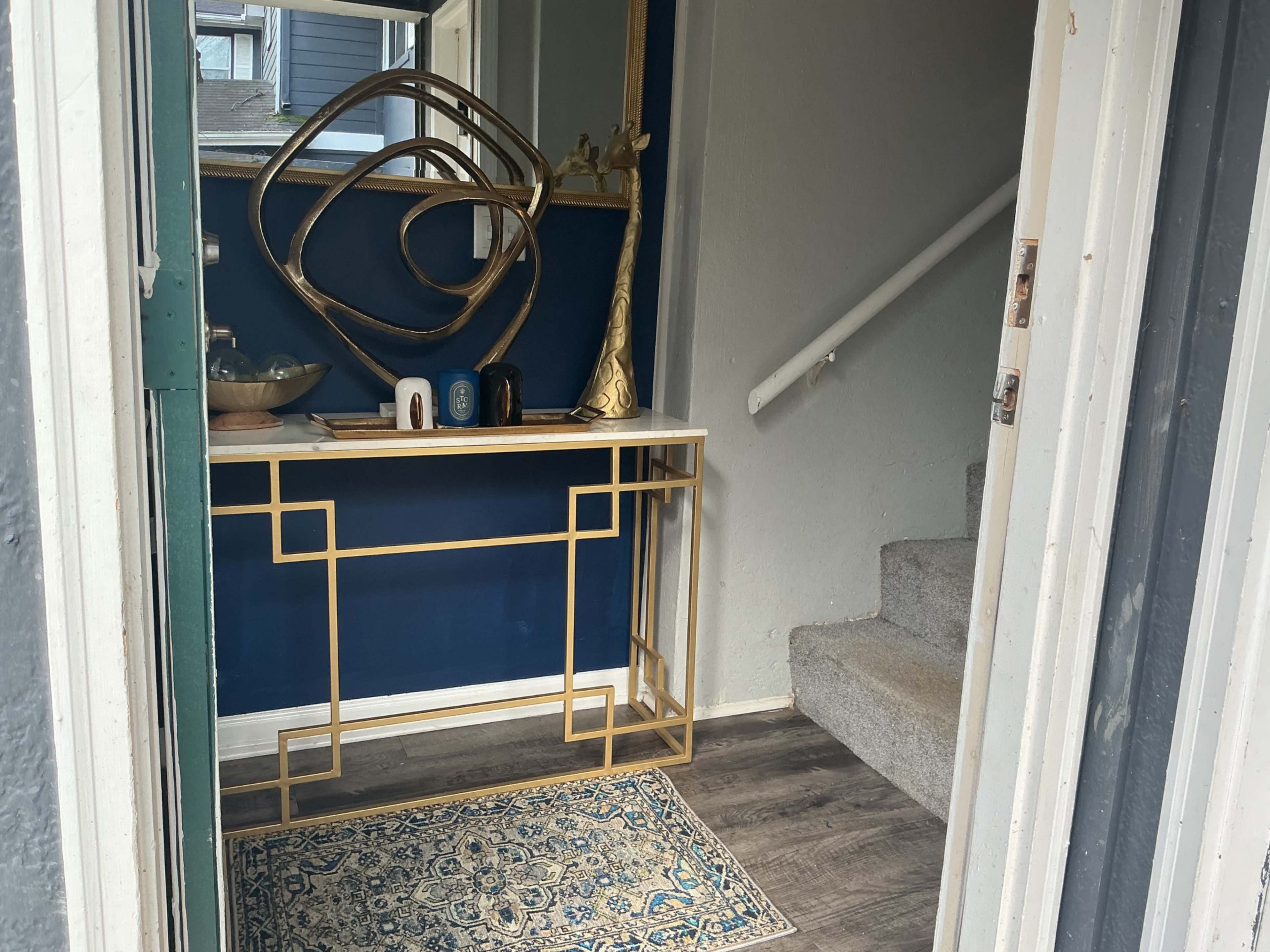 The image shows a small entryway with a decorative console table, a mirror, and a staircase leading to an upper level.