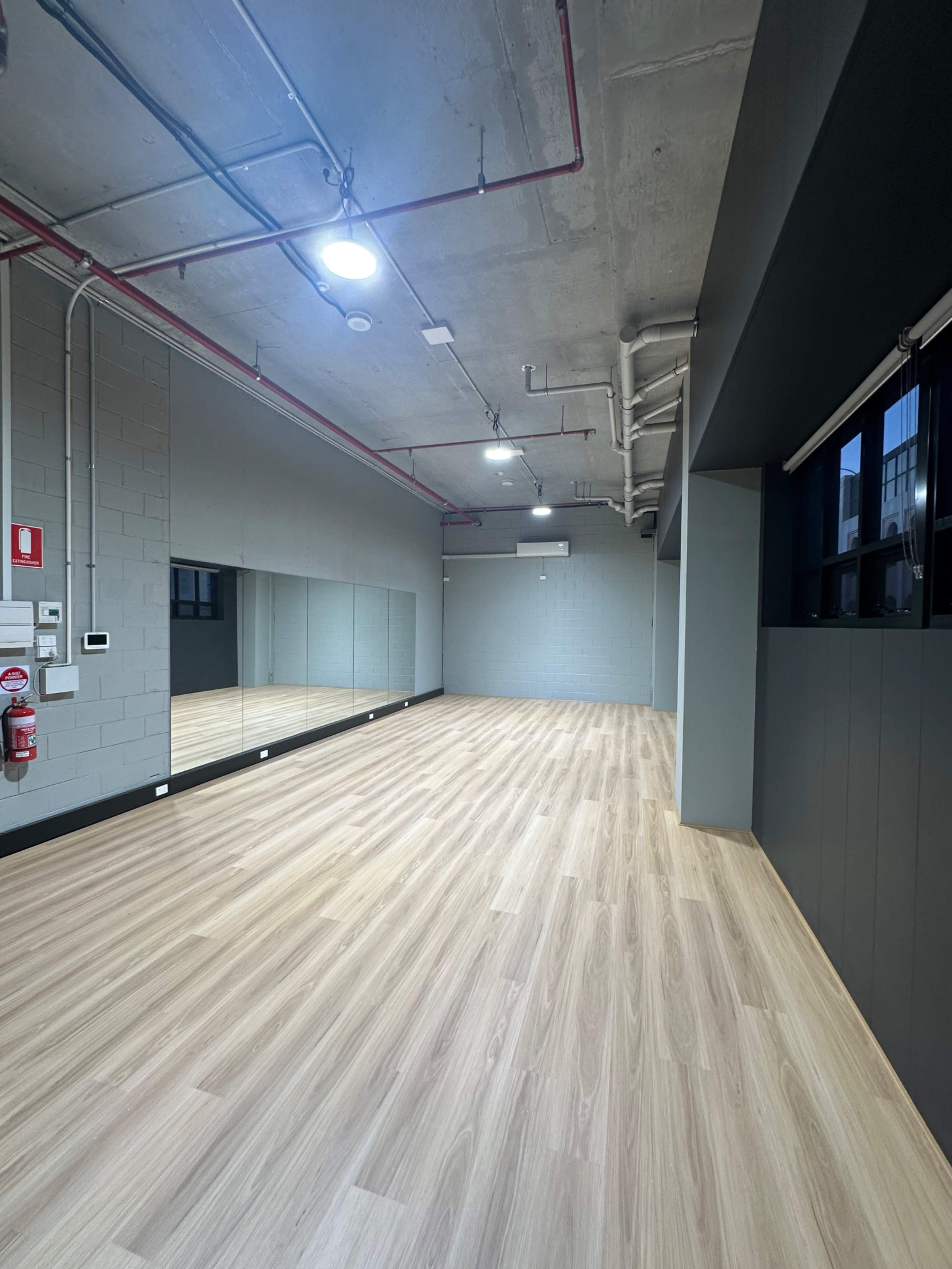 The image shows an empty studio space with wooden flooring, exposed ceiling pipes, and a large mirrored wall on one side.