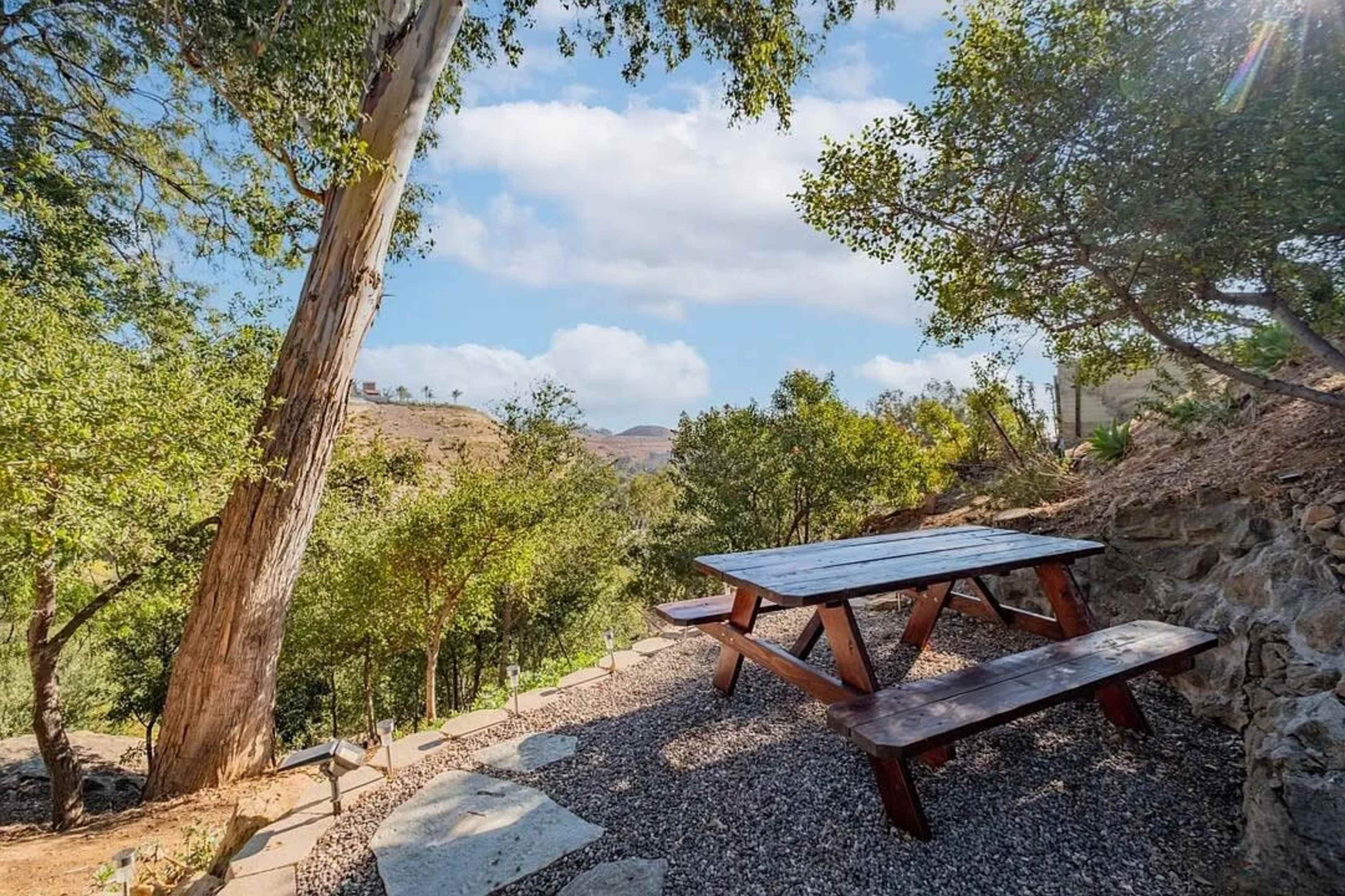 A wooden picnic table sits on gravel beneath trees, surrounded by greenery and hills under a blue sky.
