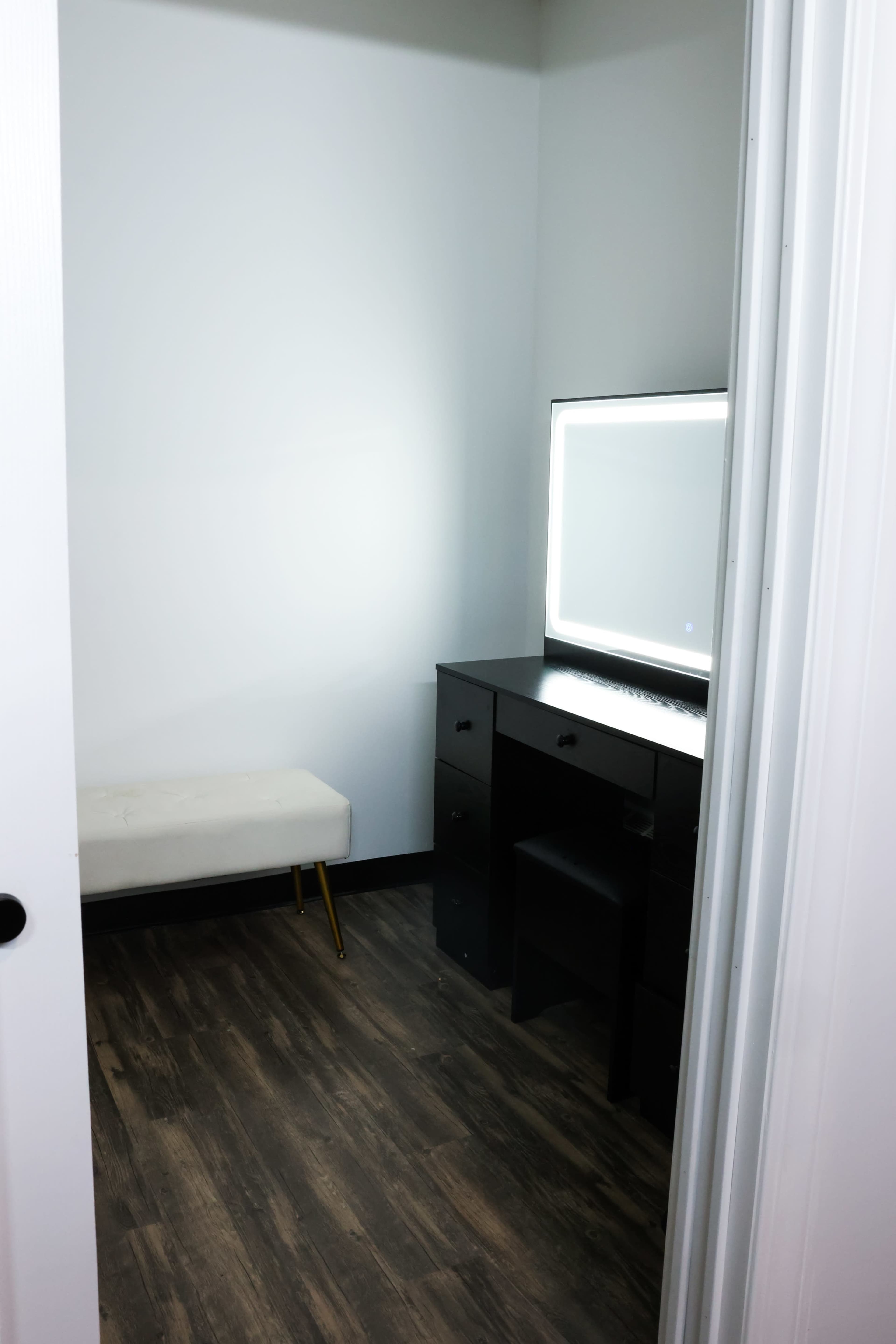 The image shows a small, well-lit room featuring a black vanity desk with a mirror and a white bench.