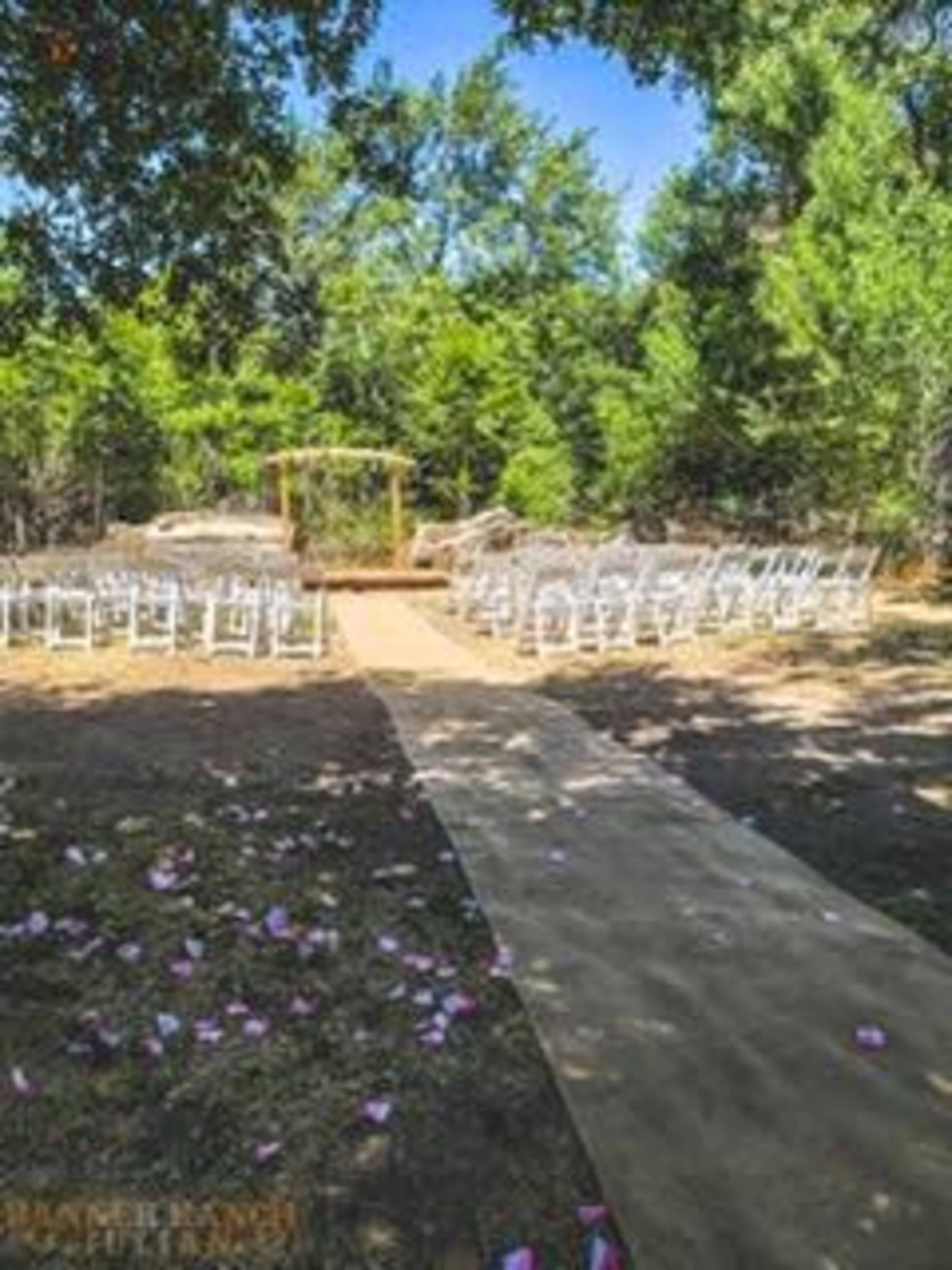 A wedding ceremony setup features white chairs arranged on either side of a pathway leading to a wooden arch, surrounded by greenery.