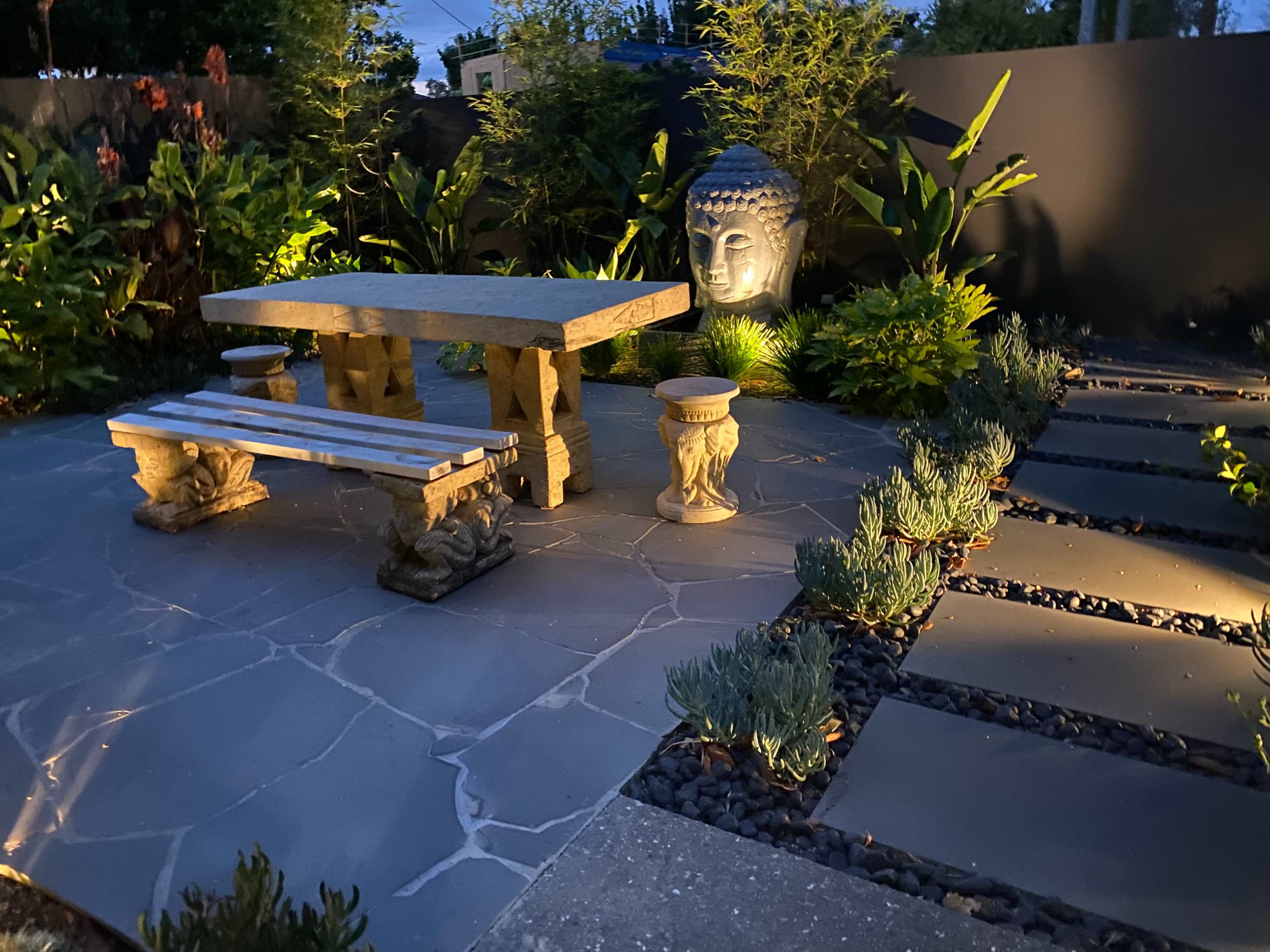 A garden scene featuring a stone table and benches surrounded by lush greenery and a statue of Buddha, illuminated by soft lighting.