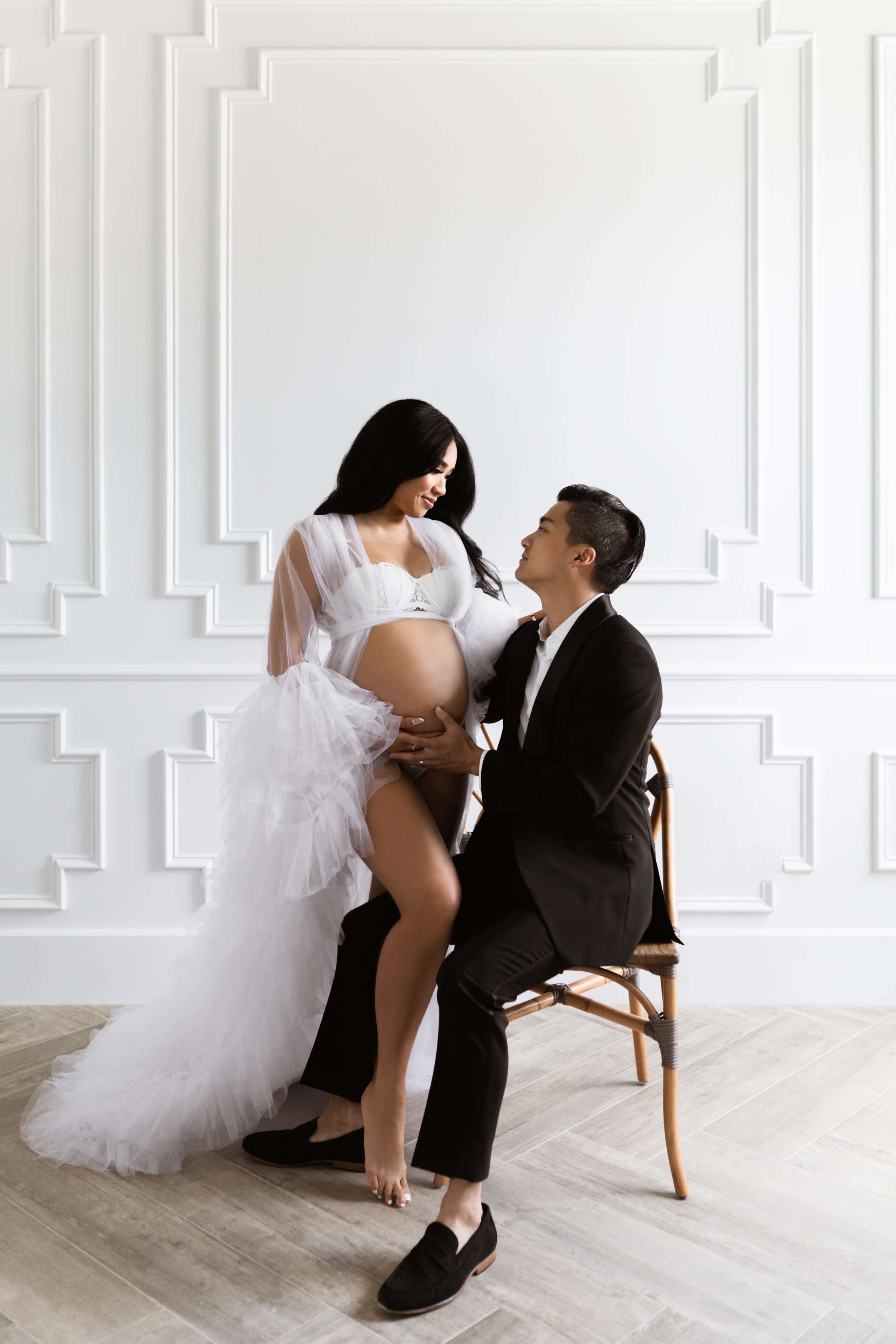 A pregnant woman in a white gown poses while sitting next to a man in a suit, both gazing at each other in a minimalist, well-lit room.