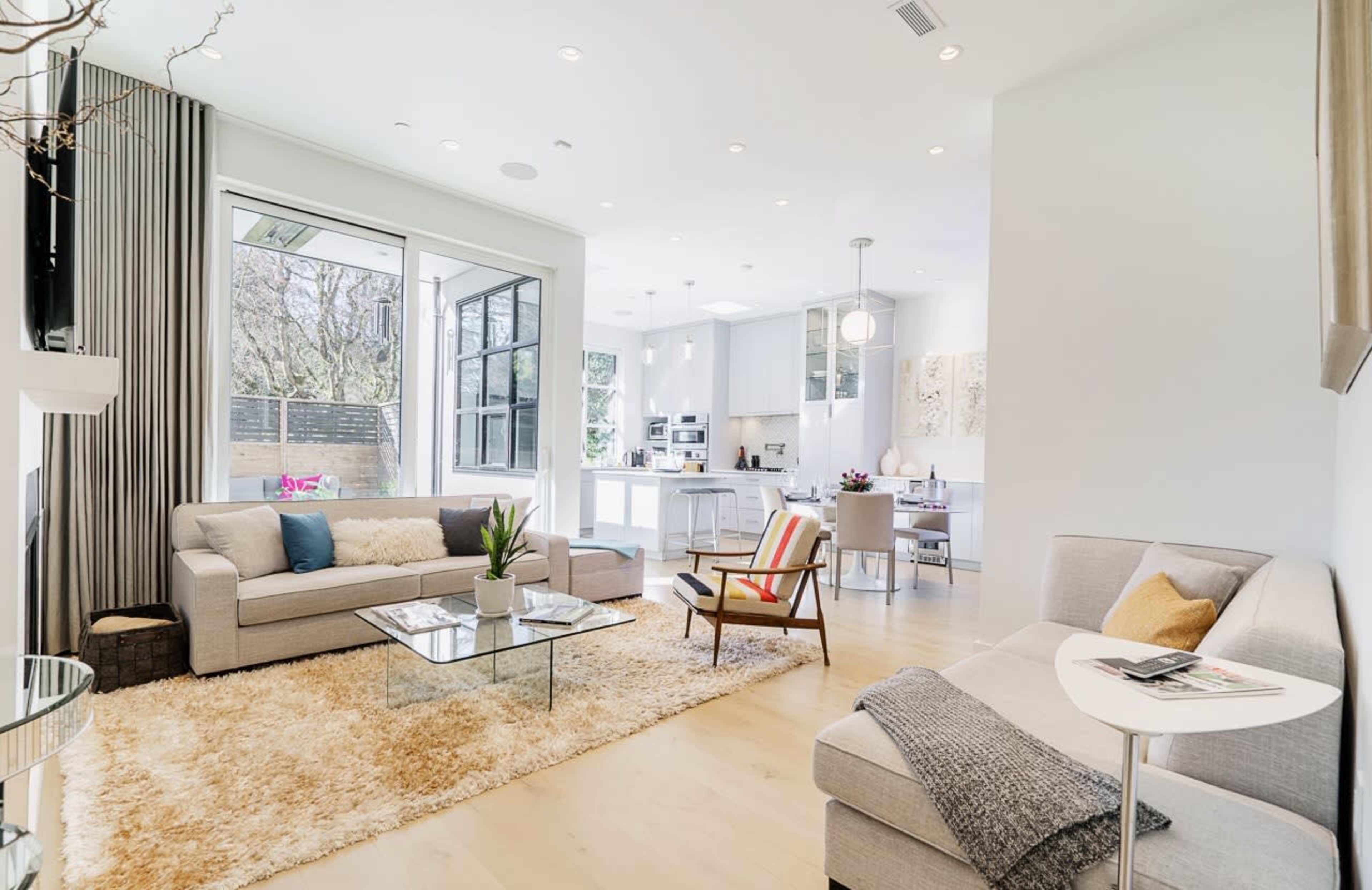 A modern living room features a light gray sofa and armchair, a glass coffee table, and large windows that overlook a well-lit kitchen area.