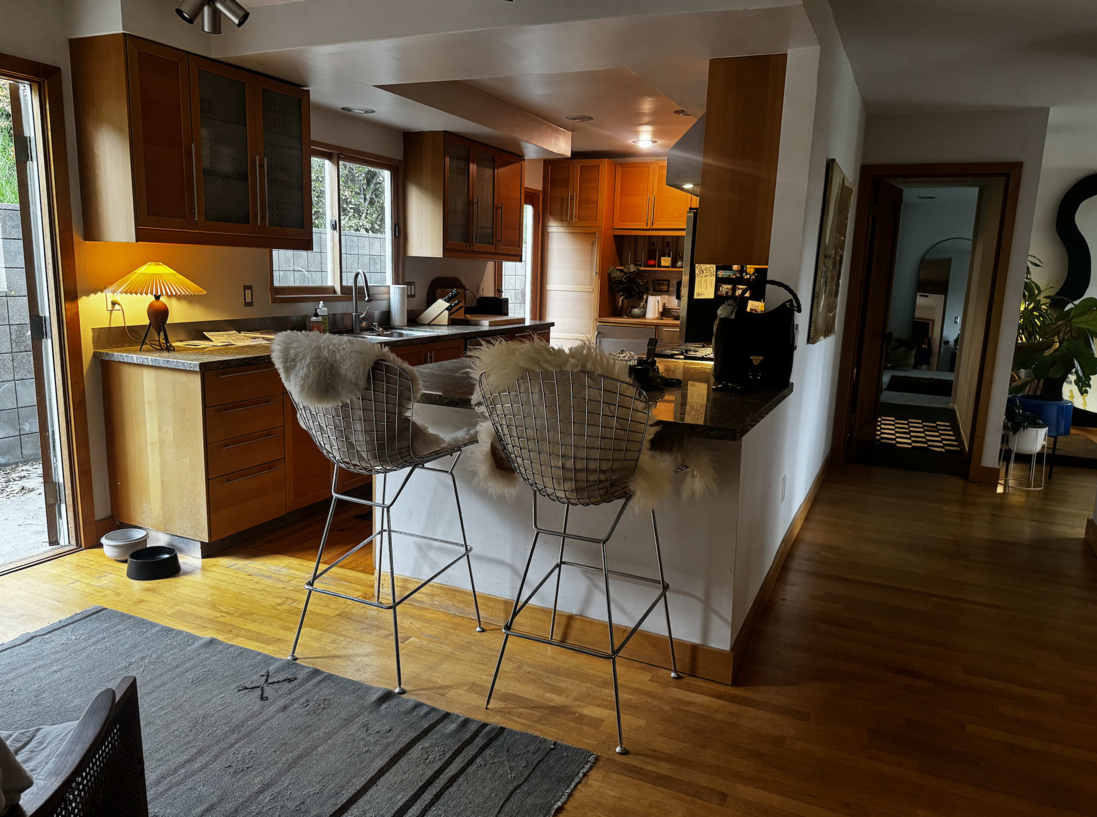 The image shows a modern kitchen with wooden cabinets, a central island with bar stools, and an open layout leading to a living space.