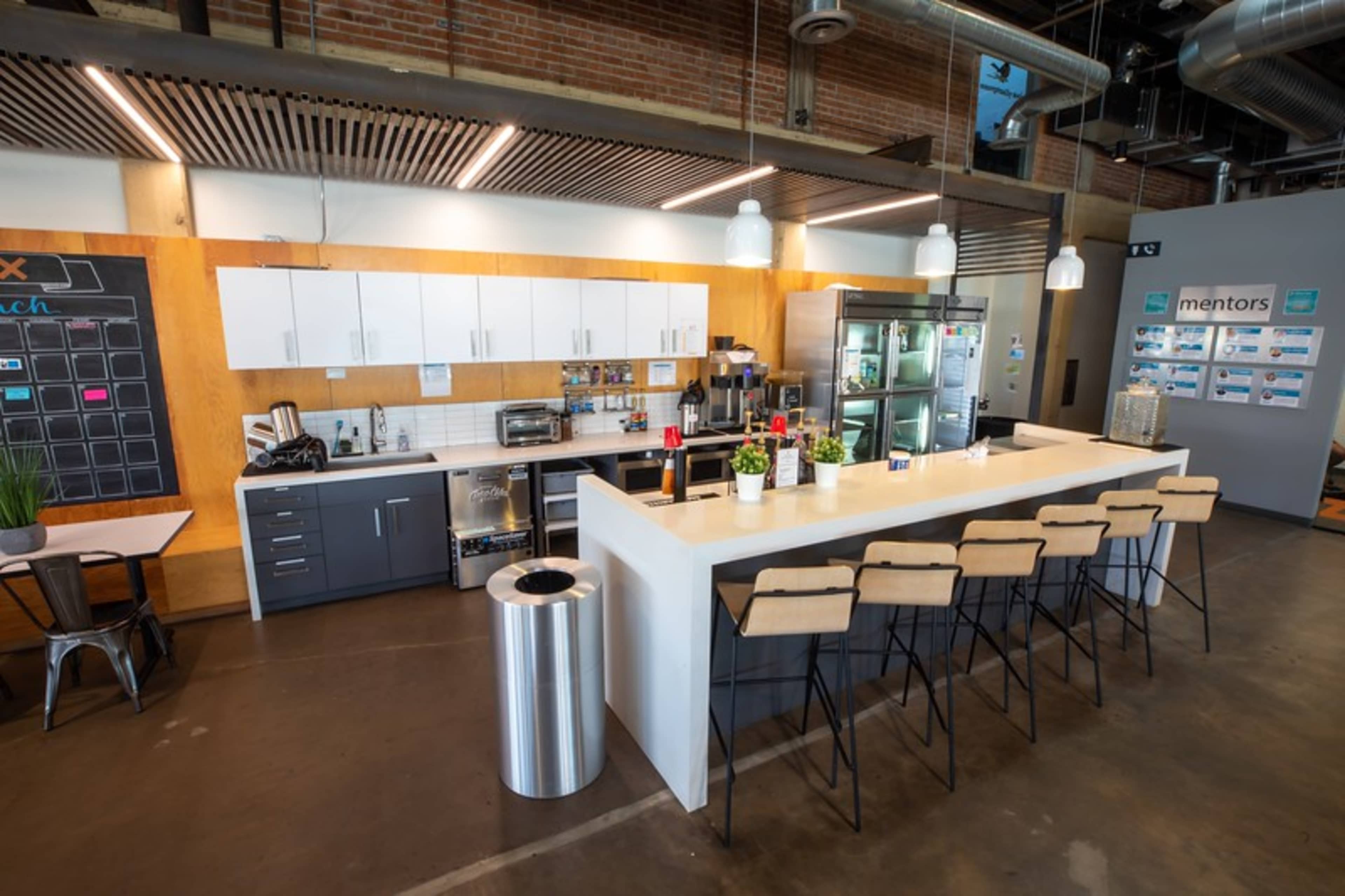 A modern kitchen area with a long white countertop, barstools, and various appliances, set against a backdrop of exposed brick and metal fixtures.