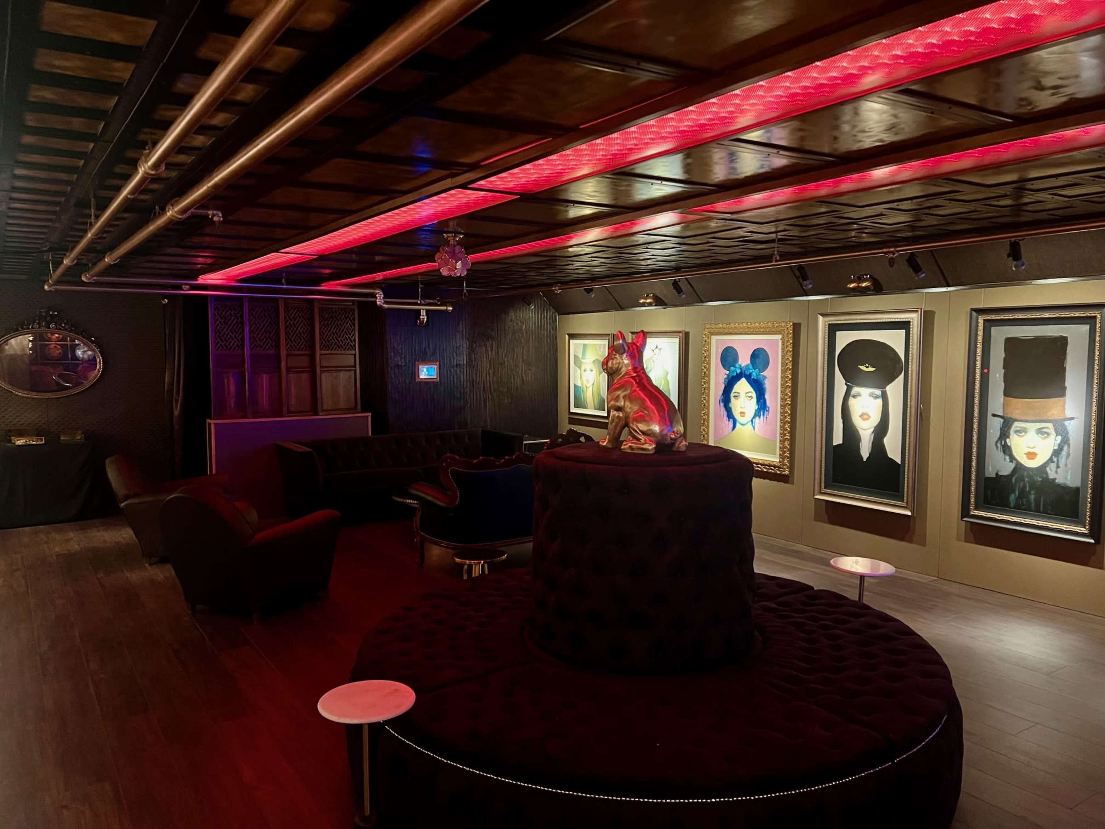 The image shows a dimly lit lounge with black velvet seating, illuminated by red lights, featuring framed artwork on the walls and a central circular seating arrangement.
