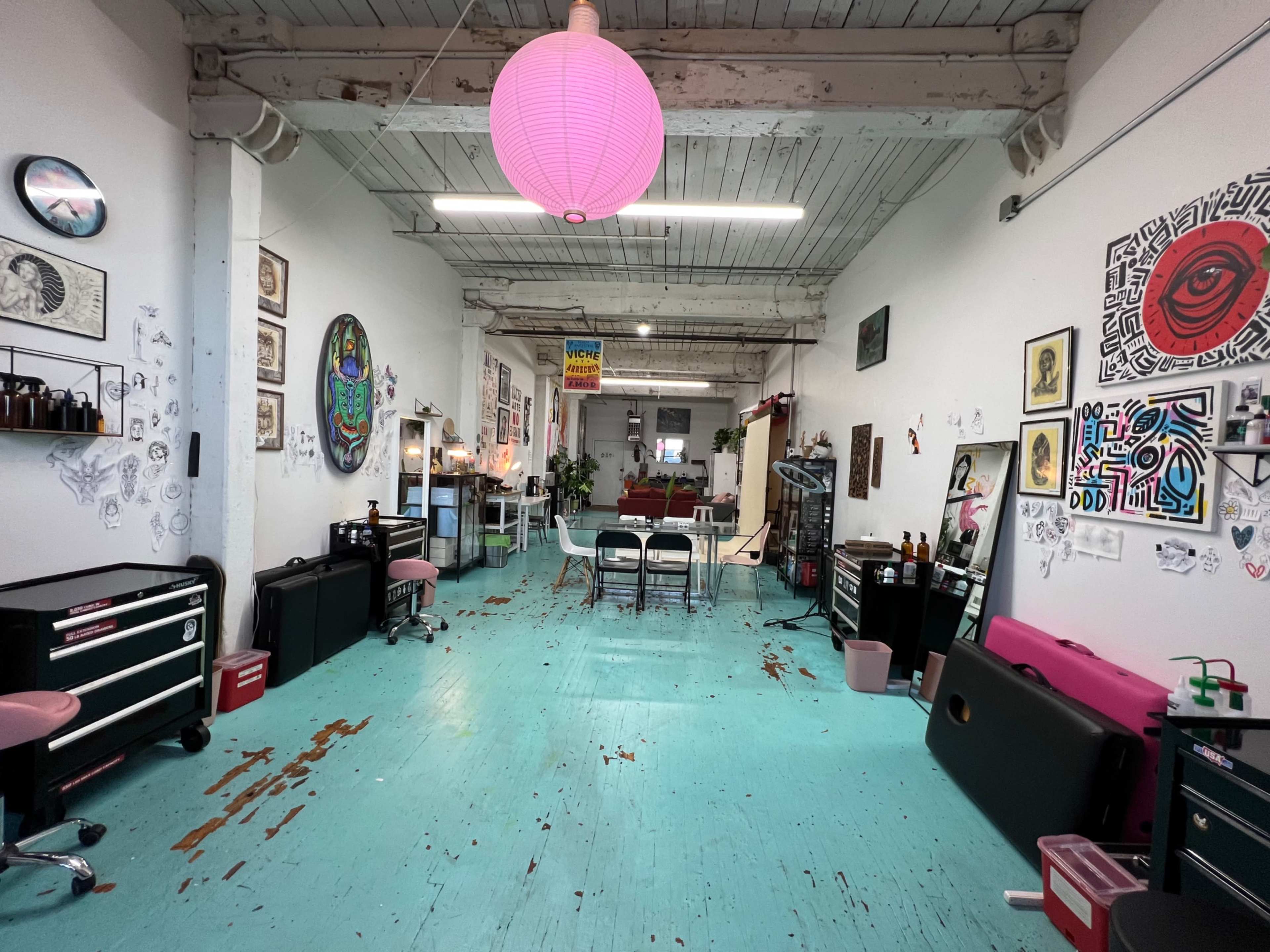 A spacious, open studio featuring teal floors, various artworks on the walls, a pink paper lantern hanging from the ceiling, and furniture arranged for creative work.