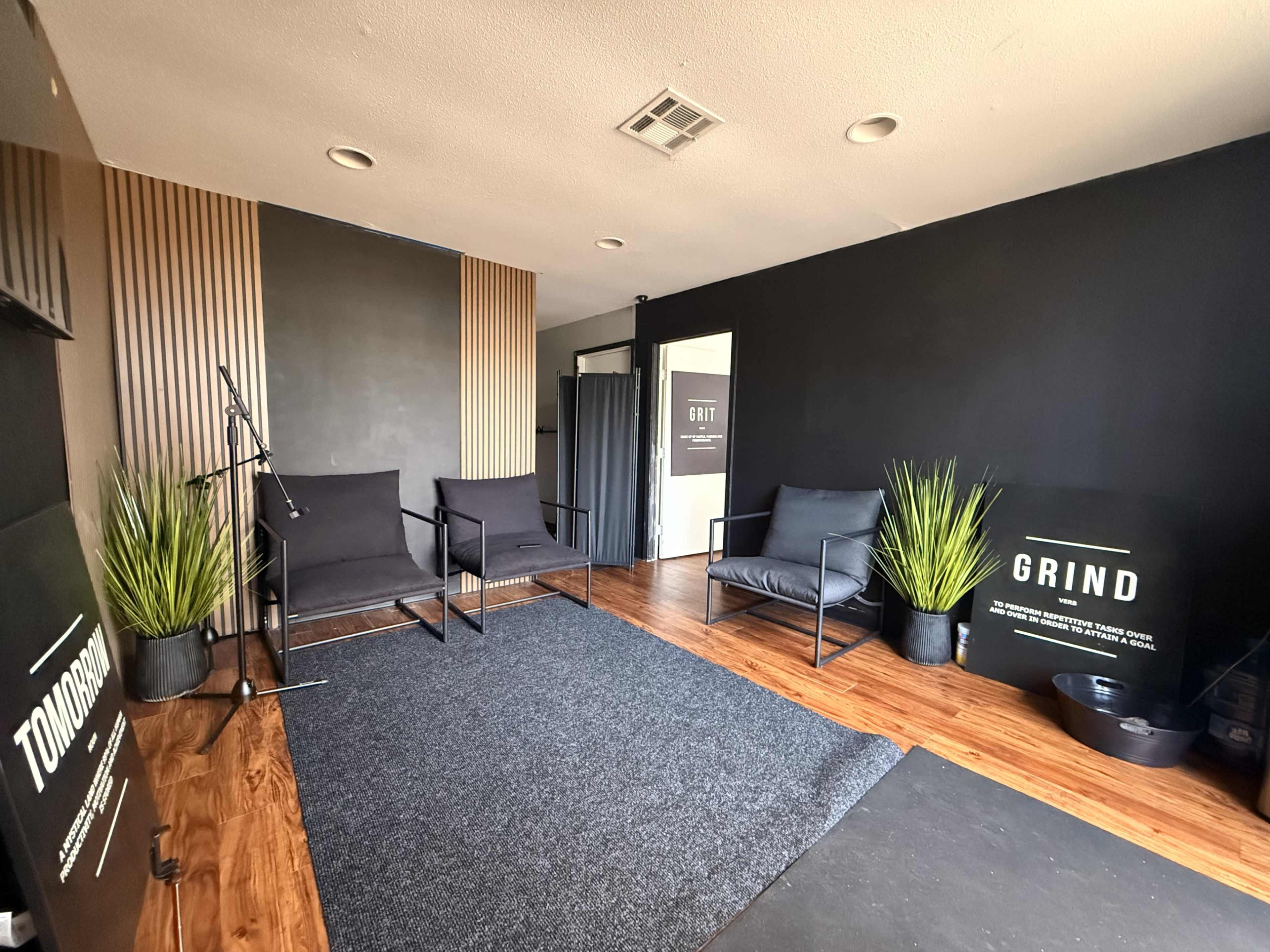 The image shows a modern, minimalistic room with black walls, a wooden floor, comfortable seating arrangements, and decorative plants.