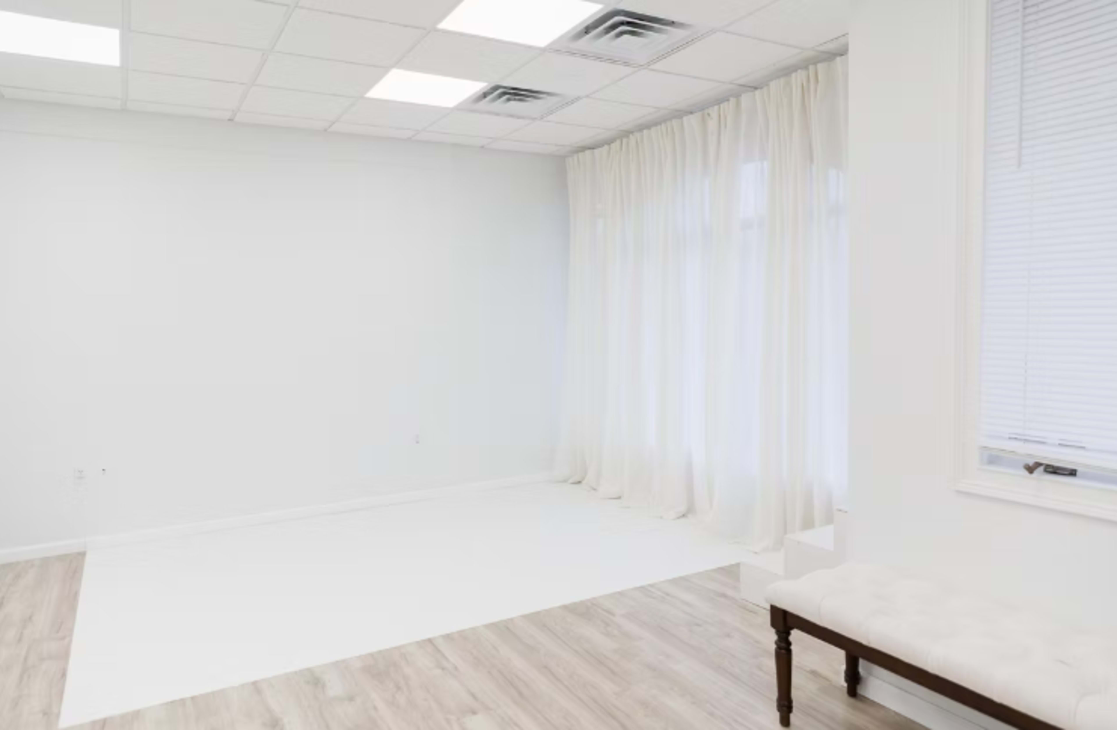 The image shows a spacious, minimalistic room with white walls, a light wooden floor, and large curtains covering a window.