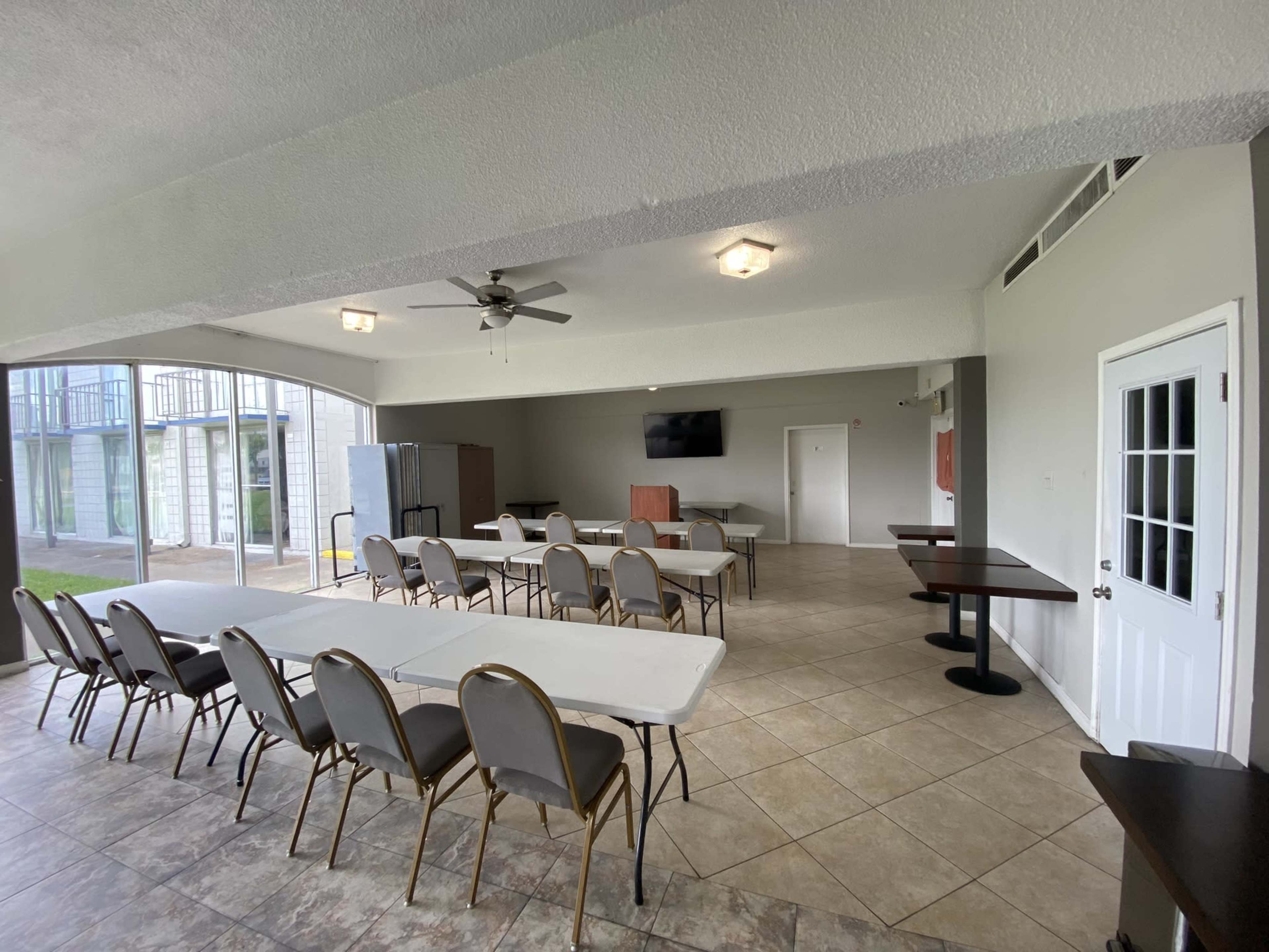 A spacious indoor room with tables and chairs arranged for a meeting, featuring a ceiling fan and a large screen on the wall.