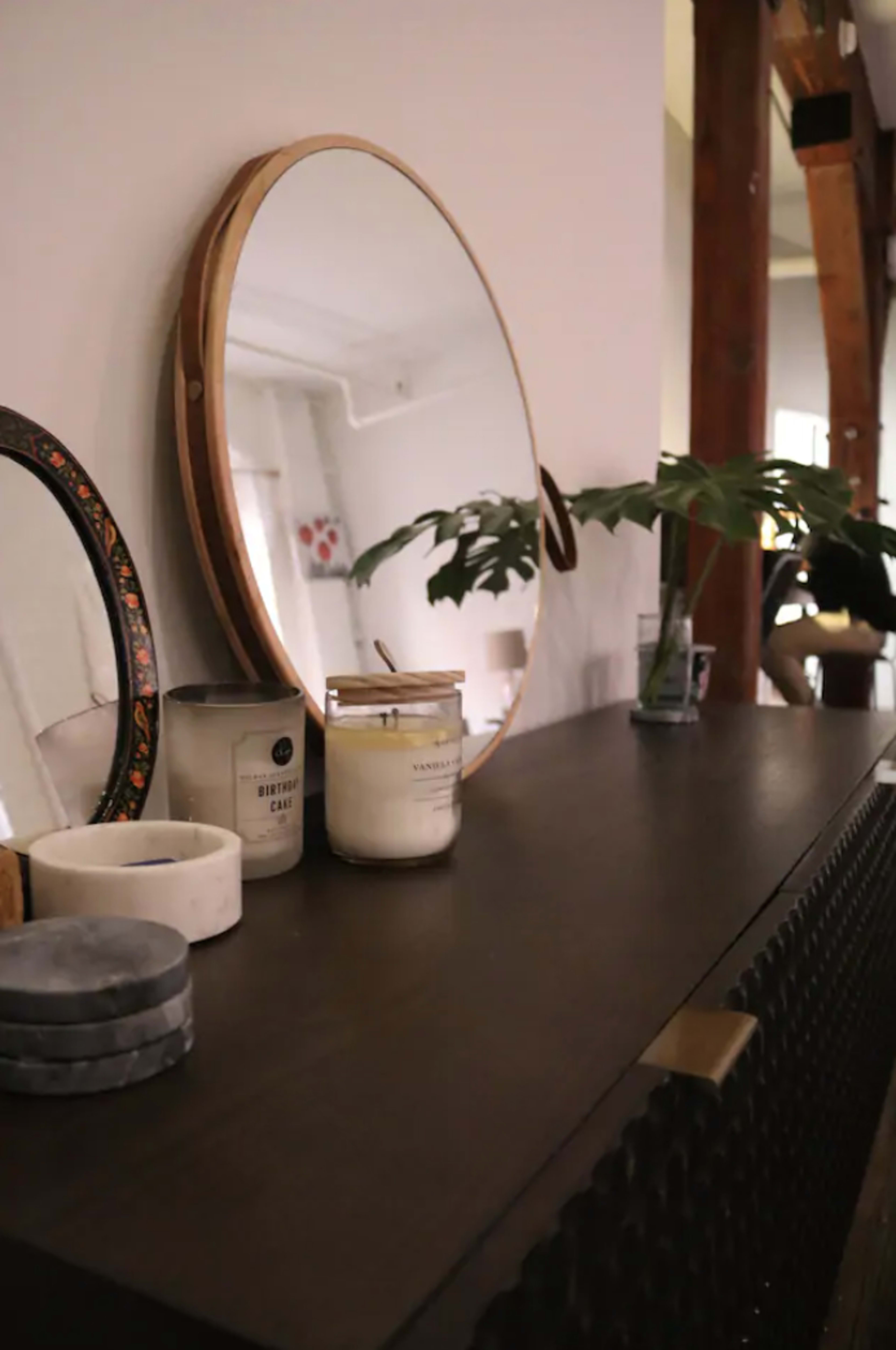A dark wooden sideboard displays a round mirror, a candle, and decorative items with a plant in the background.