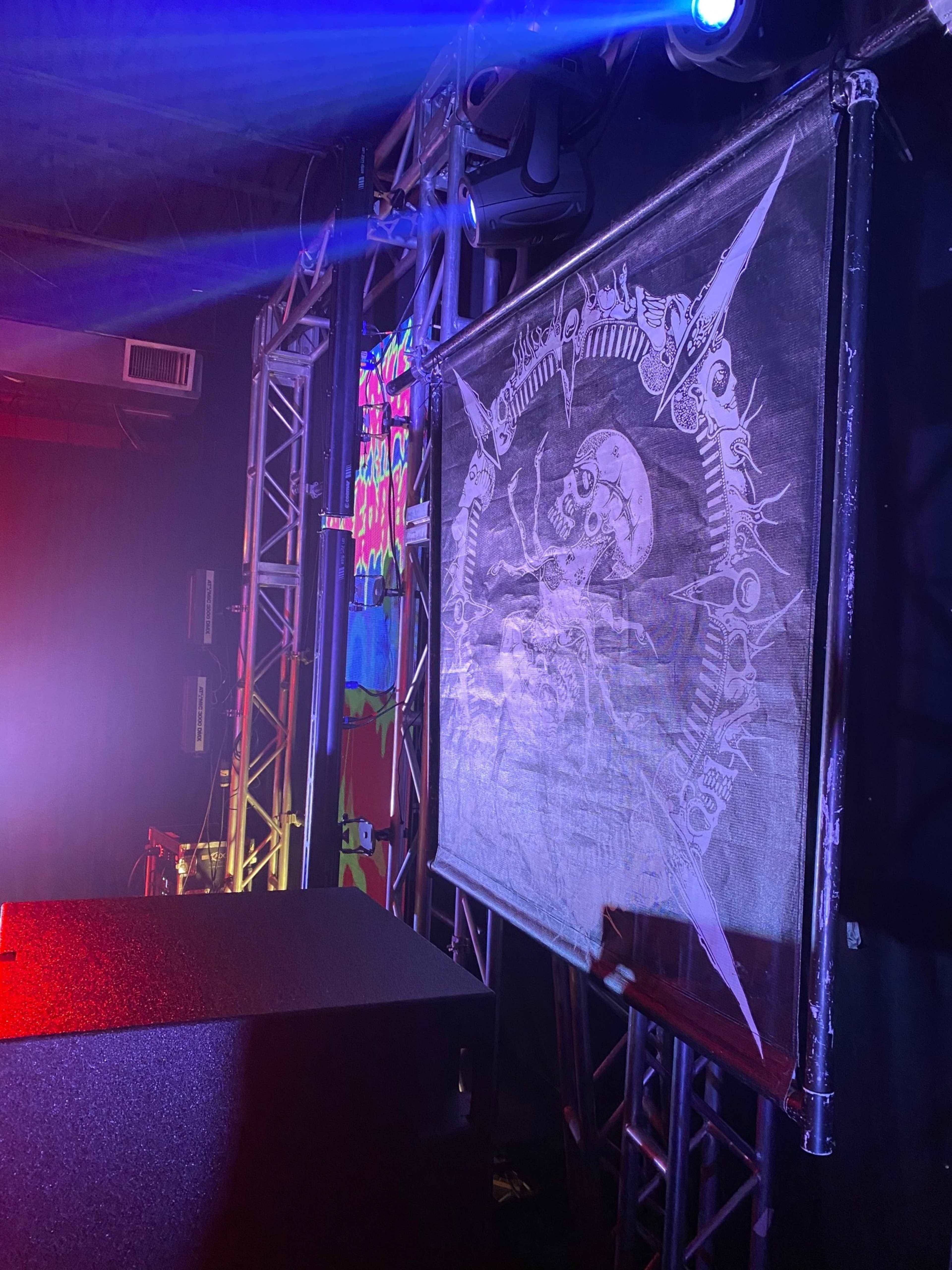 A large, detailed banner featuring a skull and intricate designs is displayed on a stage next to colorful lighting and a sound system.