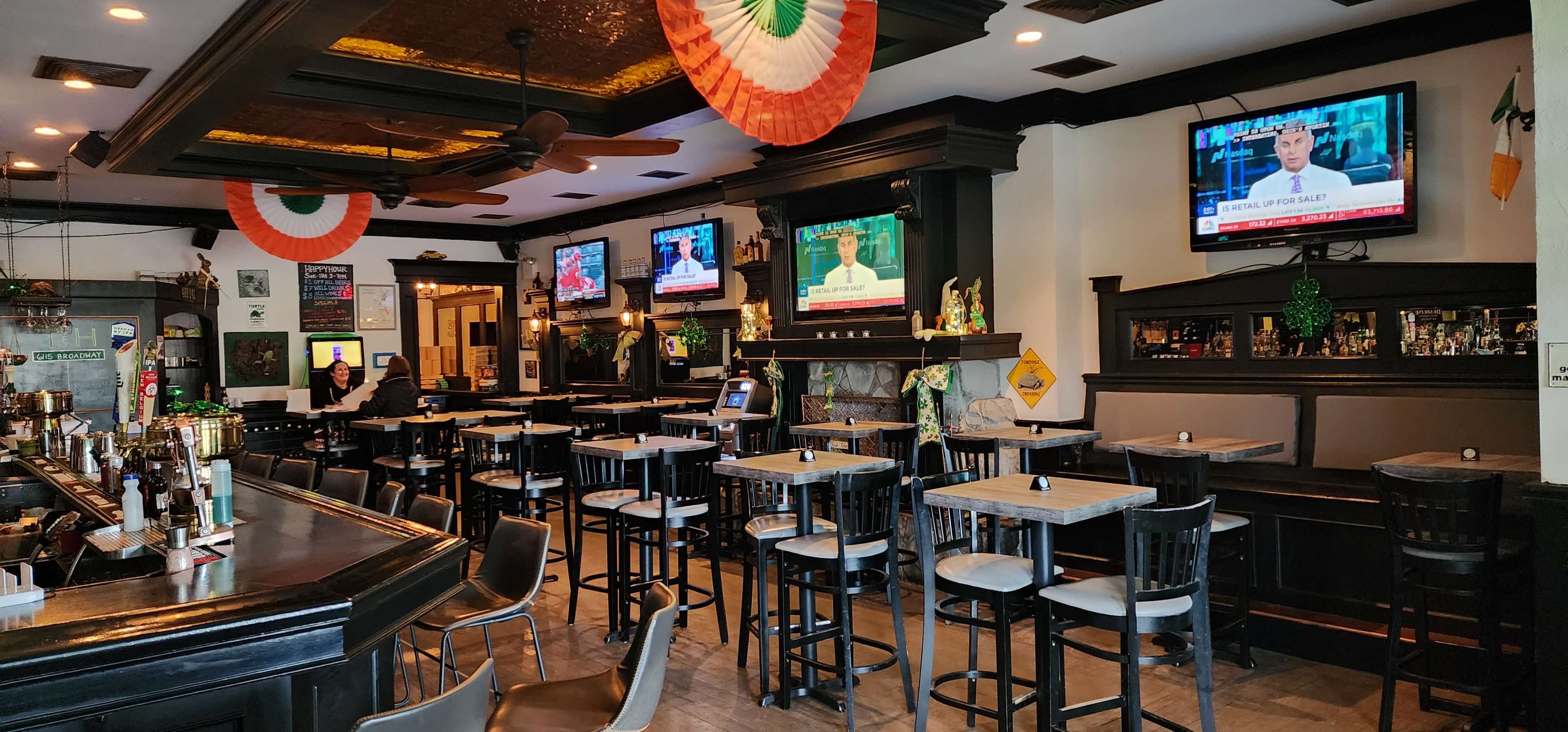 An Irish pub interior with multiple television sets displaying news, surrounded by high tables and bar stools under festive decorations.