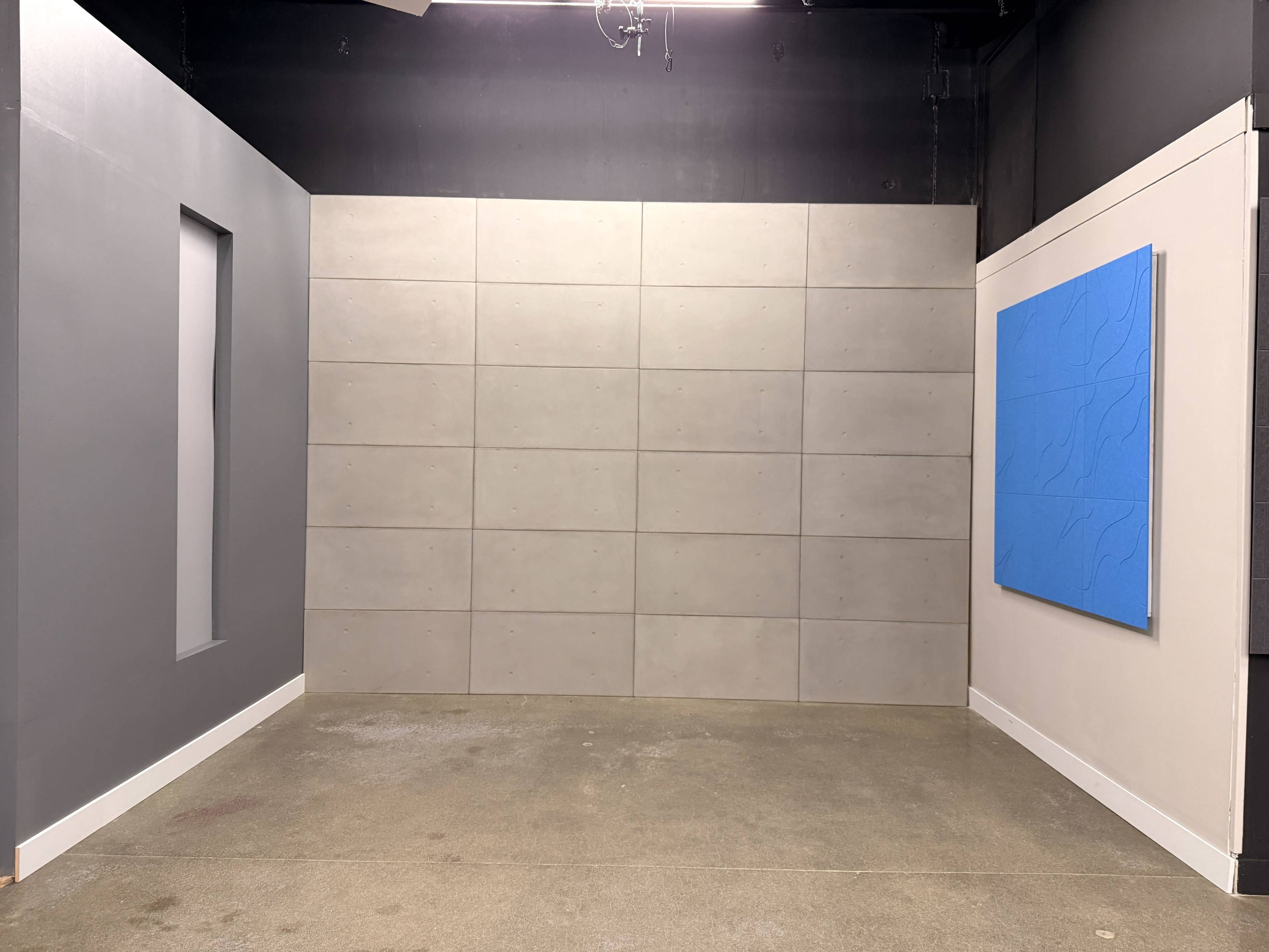 The image shows a minimalist interior setting with a gray wall featuring a vertical cutout, a large flat gray panel wall, and a blue abstract artwork mounted on a white wall.